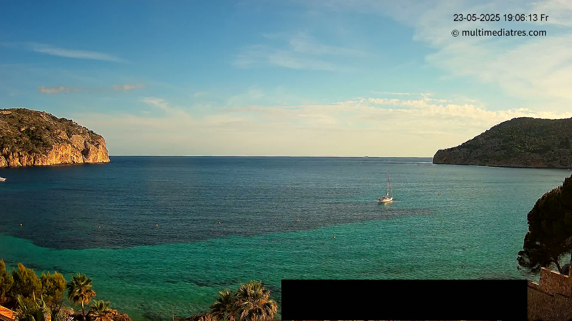 A sailboat sits in a calm, clear turquoise bay between two rocky, green hills under a mostly sunny sky.