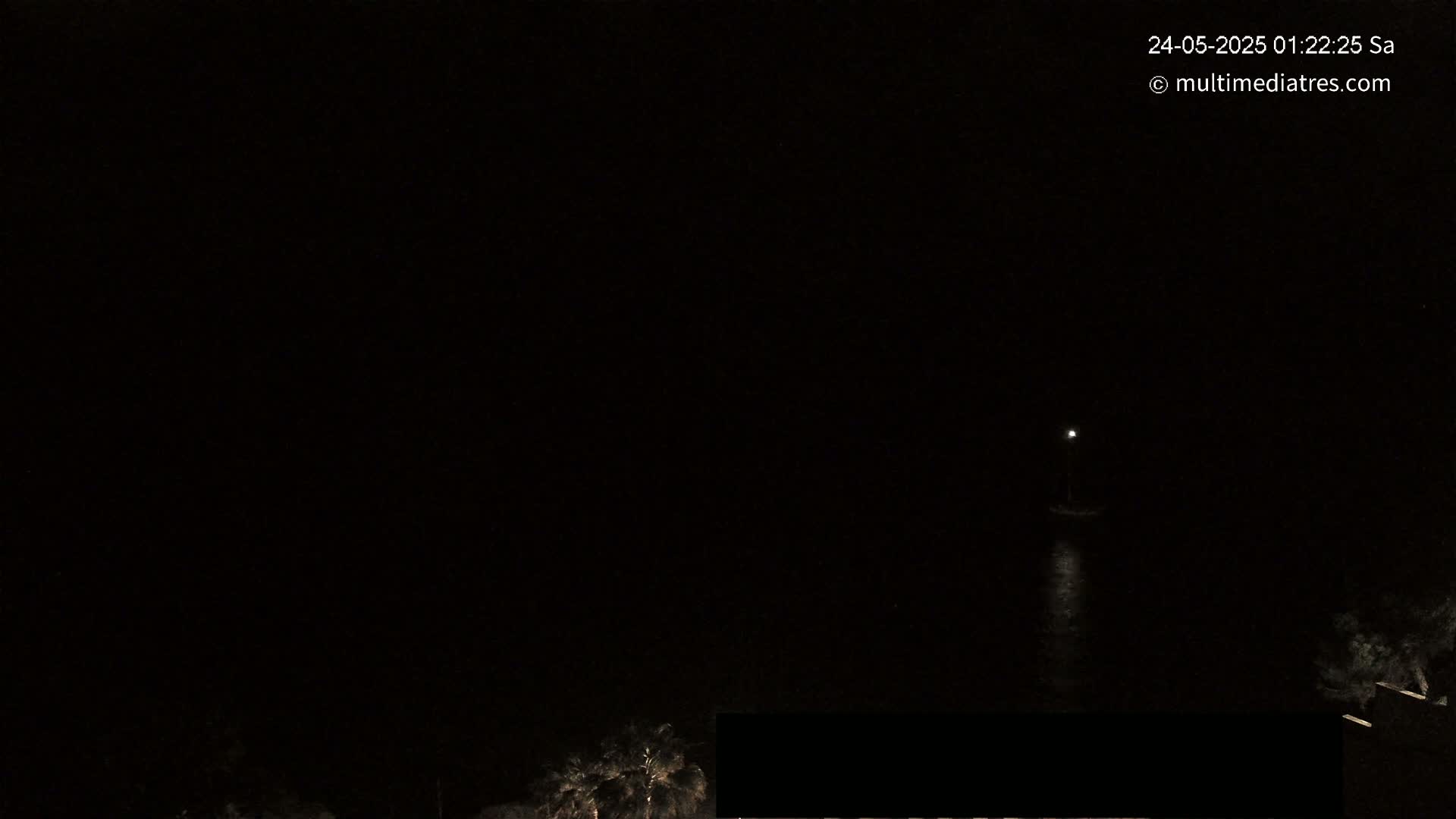 A dark nighttime scene shows a single light on the water, with some dark vegetation visible at the bottom of the frame.