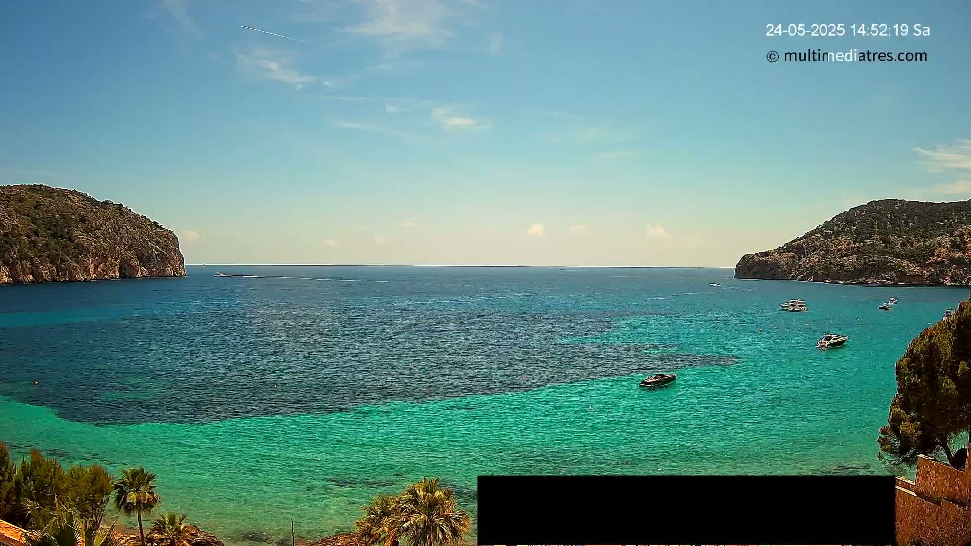 A tranquil bay with clear, turquoise water is surrounded by rocky hills under a sunny sky, with several boats scattered across the bay.