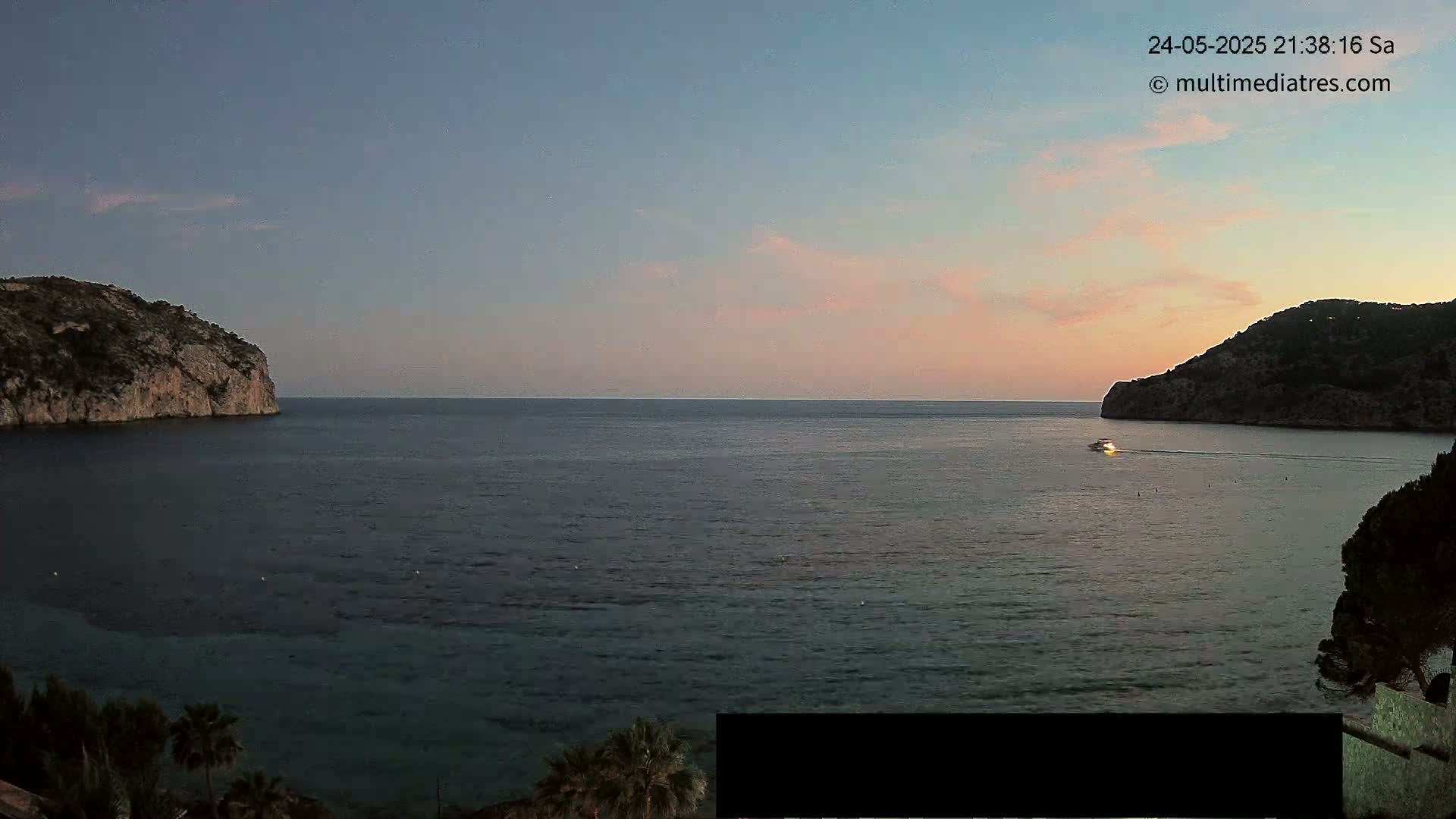A tranquil sea at dusk is nestled between two rocky hills, with a single boat visible in the distance.