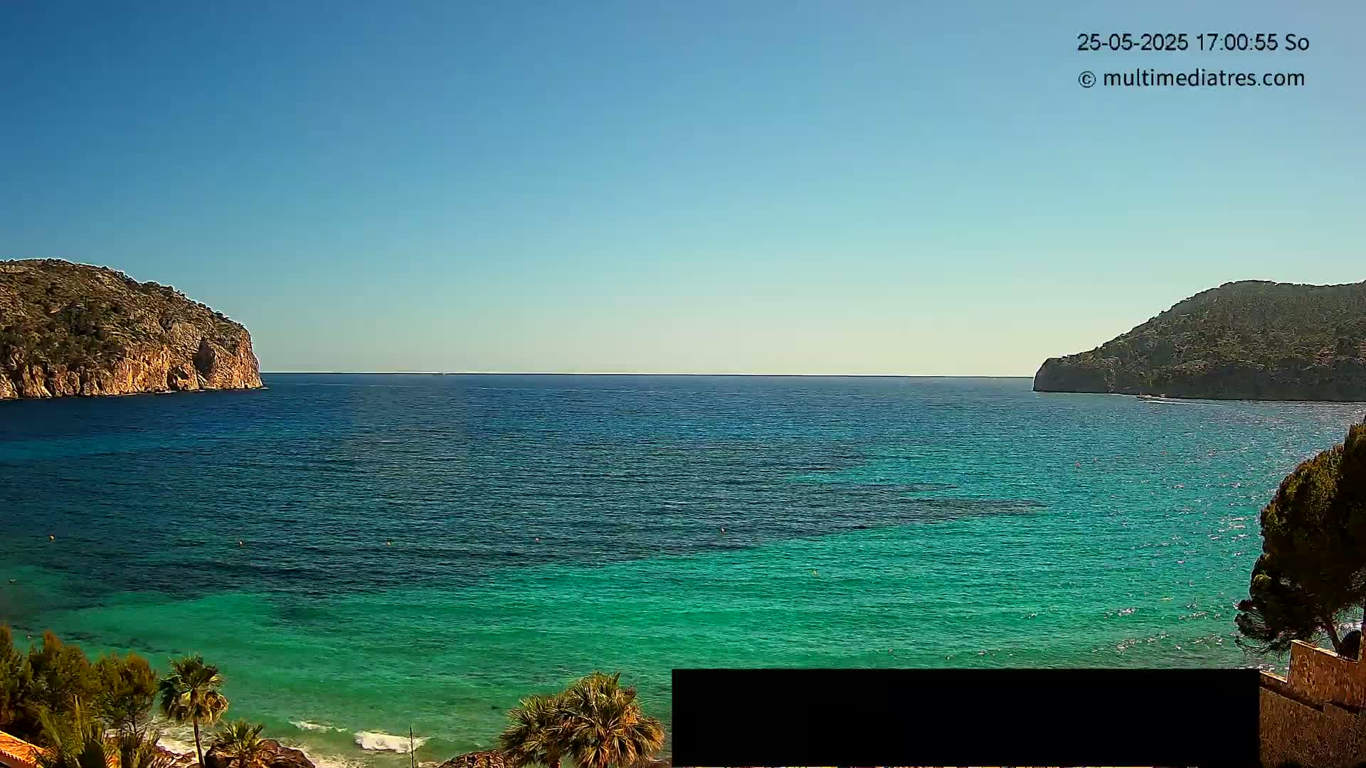 A clear, sunny day reveals a calm, turquoise bay nestled between two rocky hillsides.