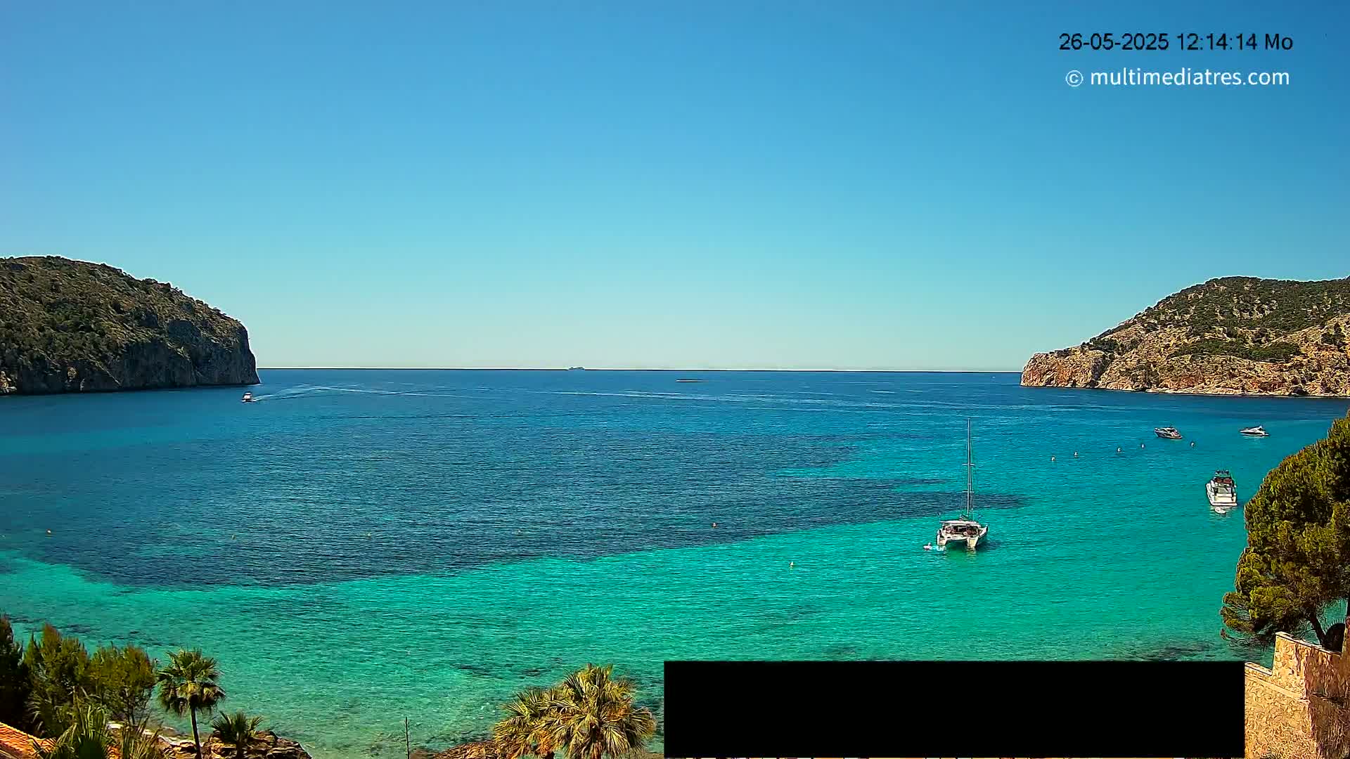 A tranquil bay with clear, turquoise water is surrounded by rocky hills under a clear blue sky.
