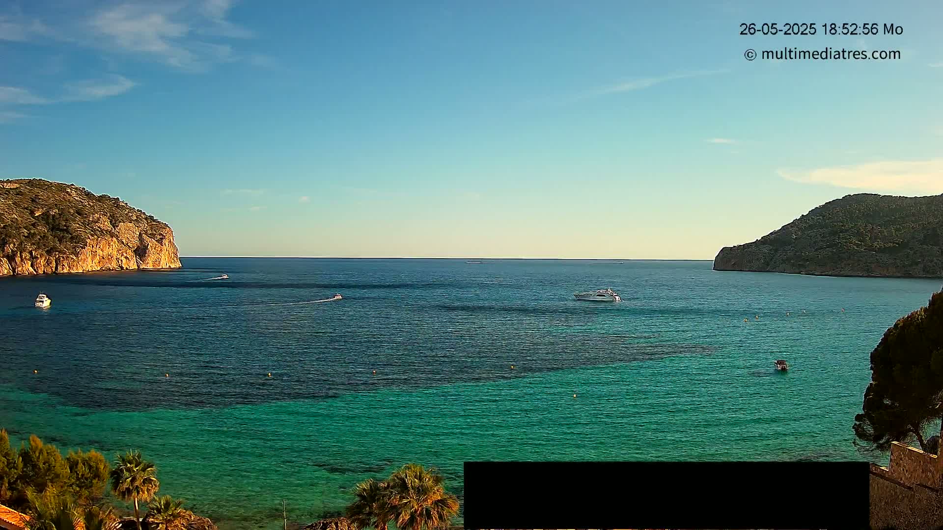A calm, sunny day shows a cove of clear, teal water surrounded by rocky hills and a few boats.
