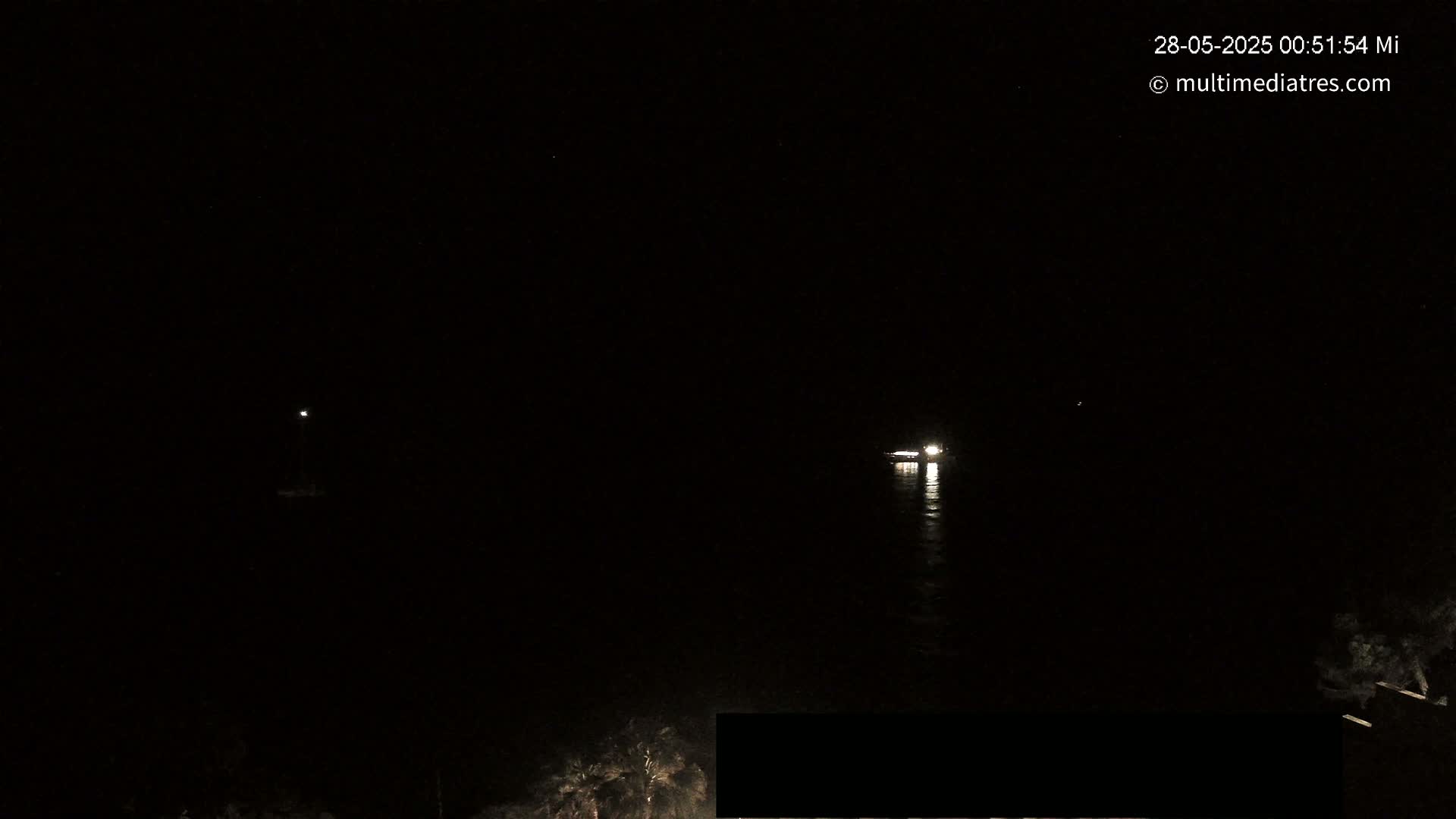 At night, two faintly lit vessels are visible on dark water, with some vegetation on the lower right.