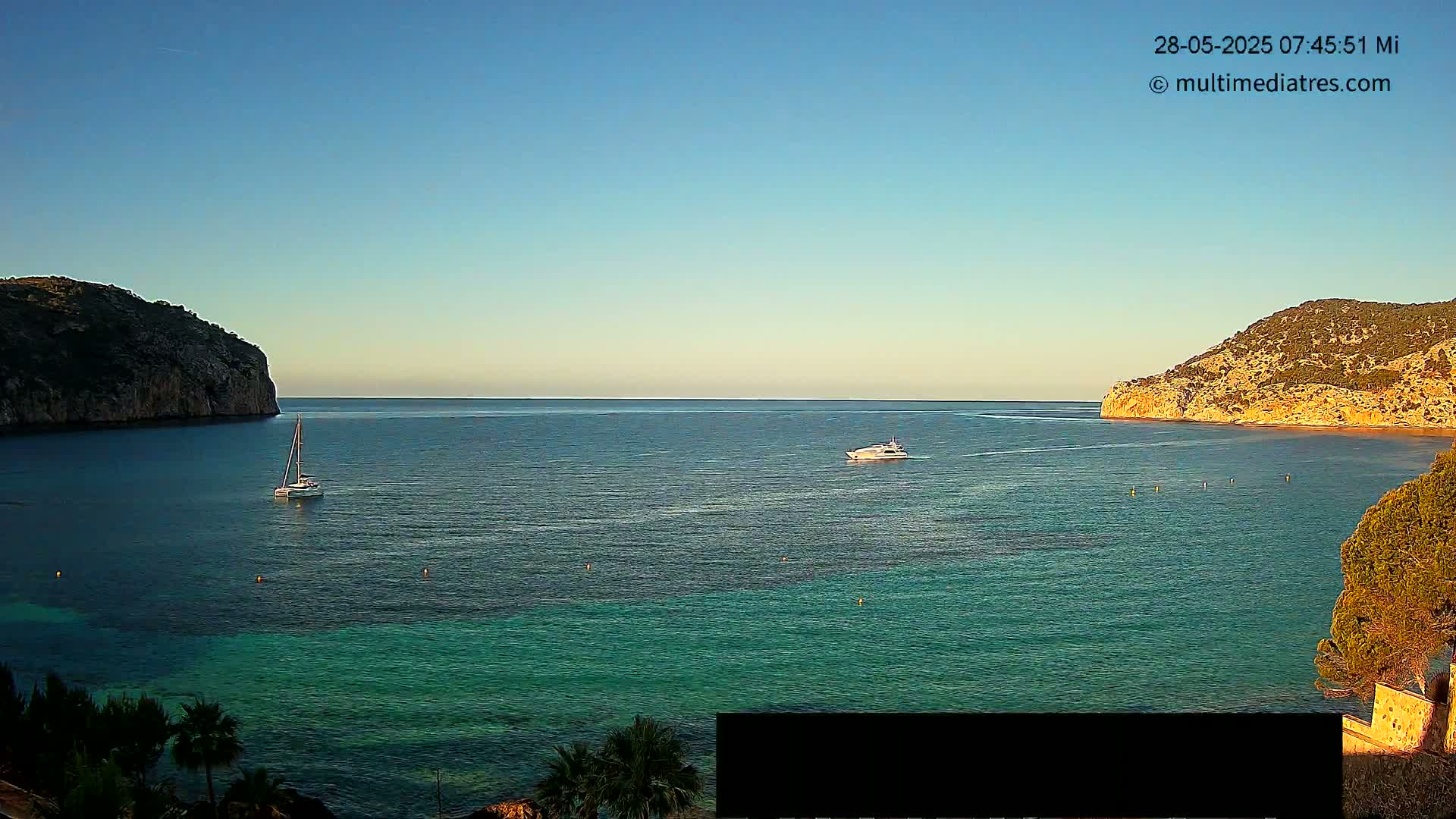A calm, clear day reveals a tranquil bay nestled between two rocky hills, with a sailboat and a motorboat visible on the turquoise water.