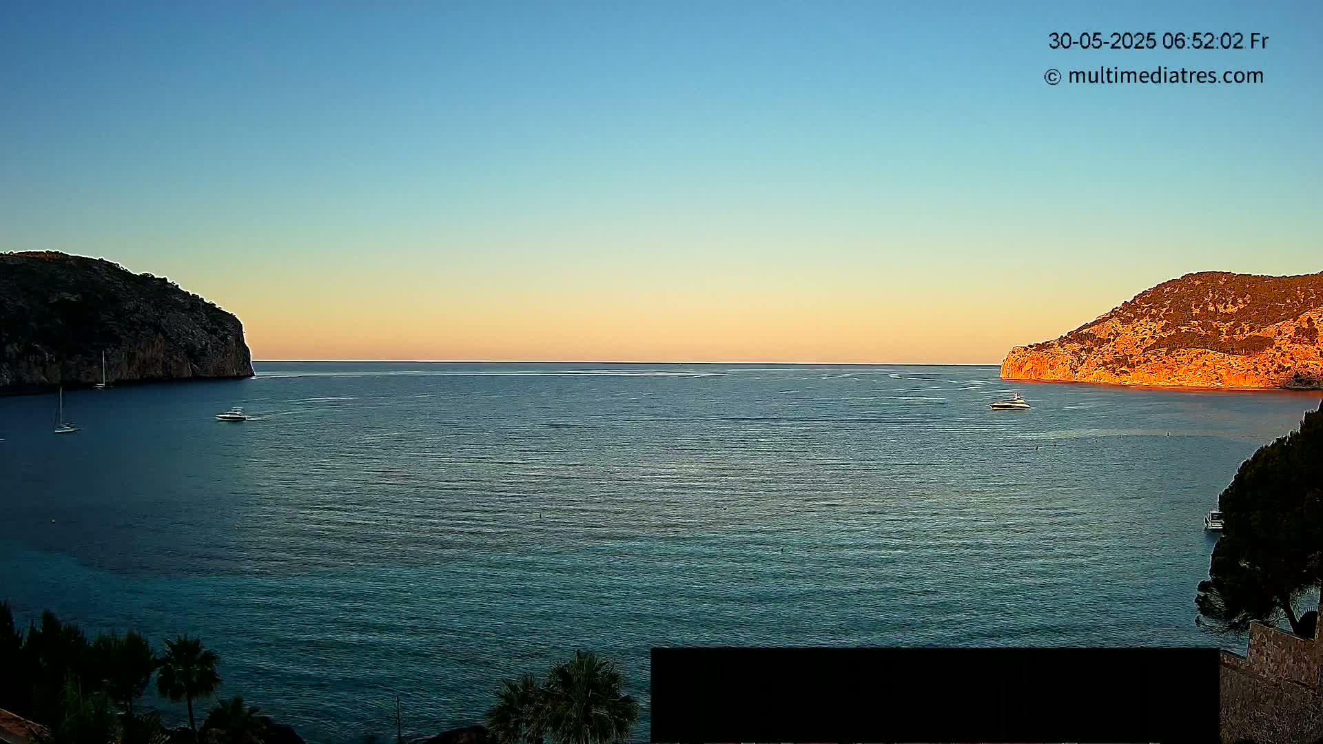 A calm sea at sunset is nestled between two rocky hills, with a few boats scattered across the water.