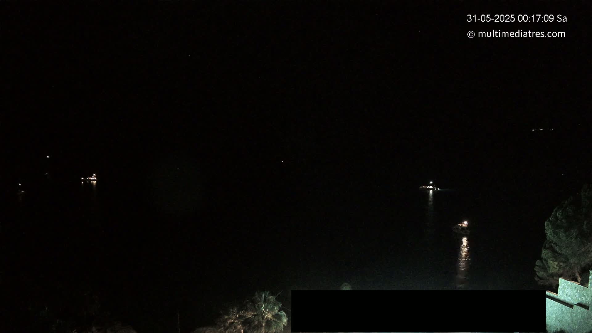 At night, several small lights are visible on the dark water, possibly from boats, with a darker shoreline and some vegetation in the bottom right corner.