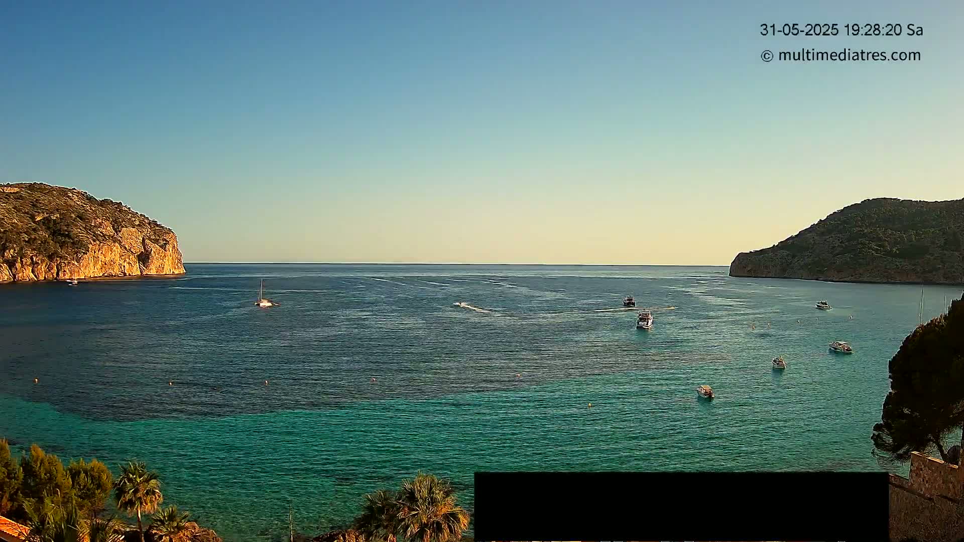 A calm, clear day reveals a tranquil bay with several small boats and sailboats, surrounded by dark-green hillsides, and the water changes from a deep blue to a vibrant teal closer to shore.