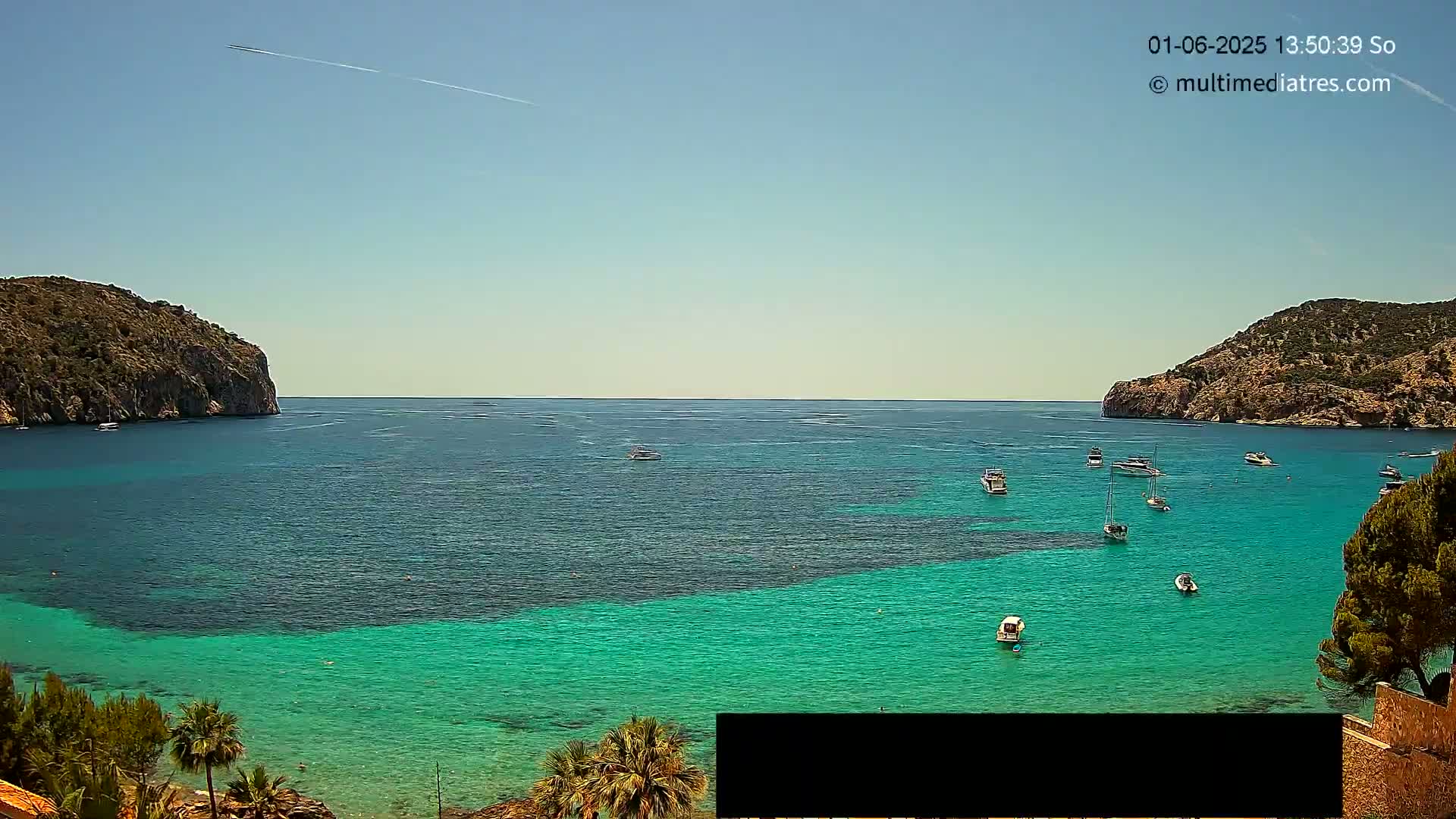 A tranquil bay with clear, turquoise water is surrounded by rocky hills under a clear, sunny sky, with several small boats scattered across the water.