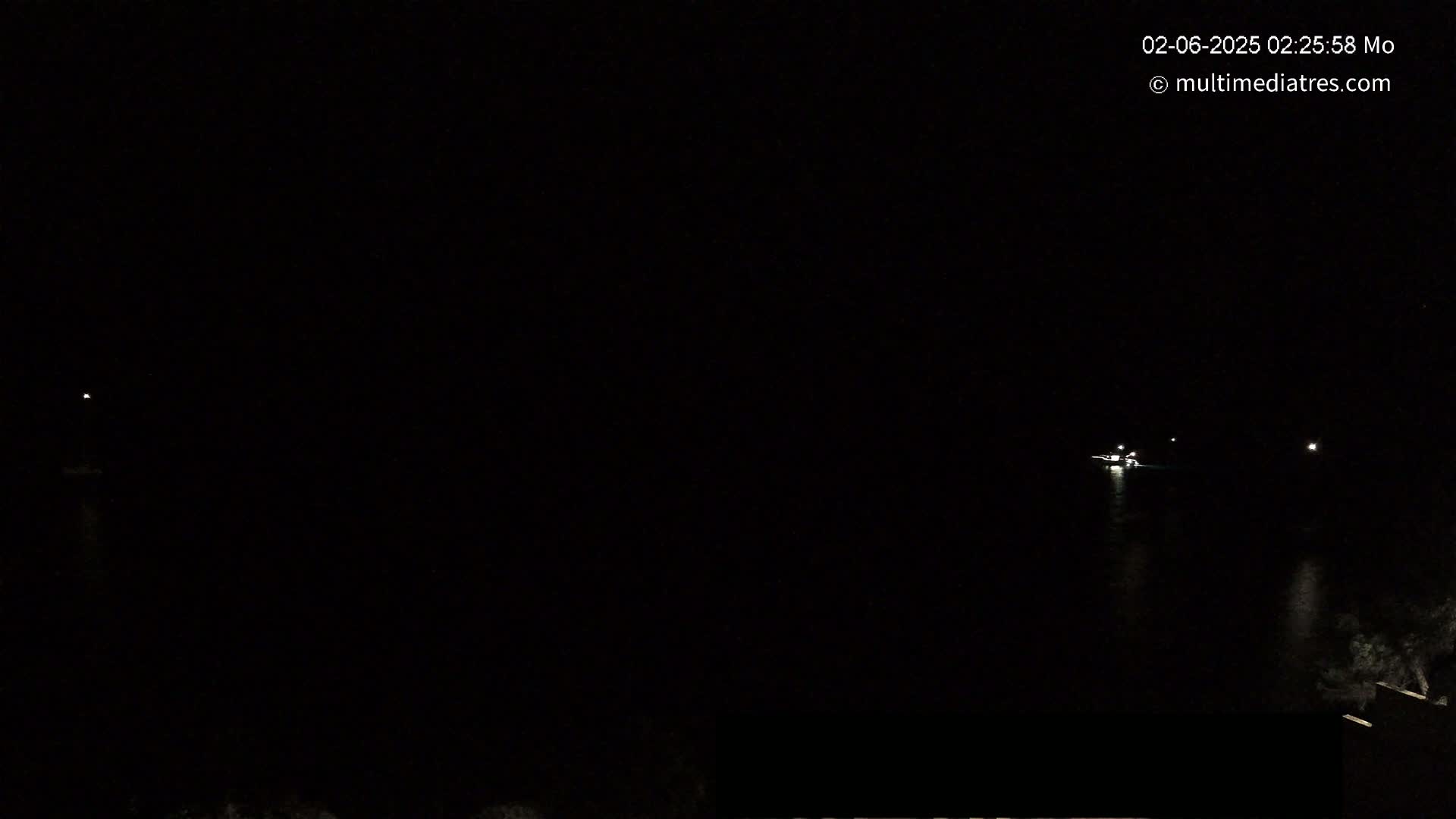A nighttime, dark image shows a body of water with a few faint lights on it and a bit of shoreline visible on the bottom right.