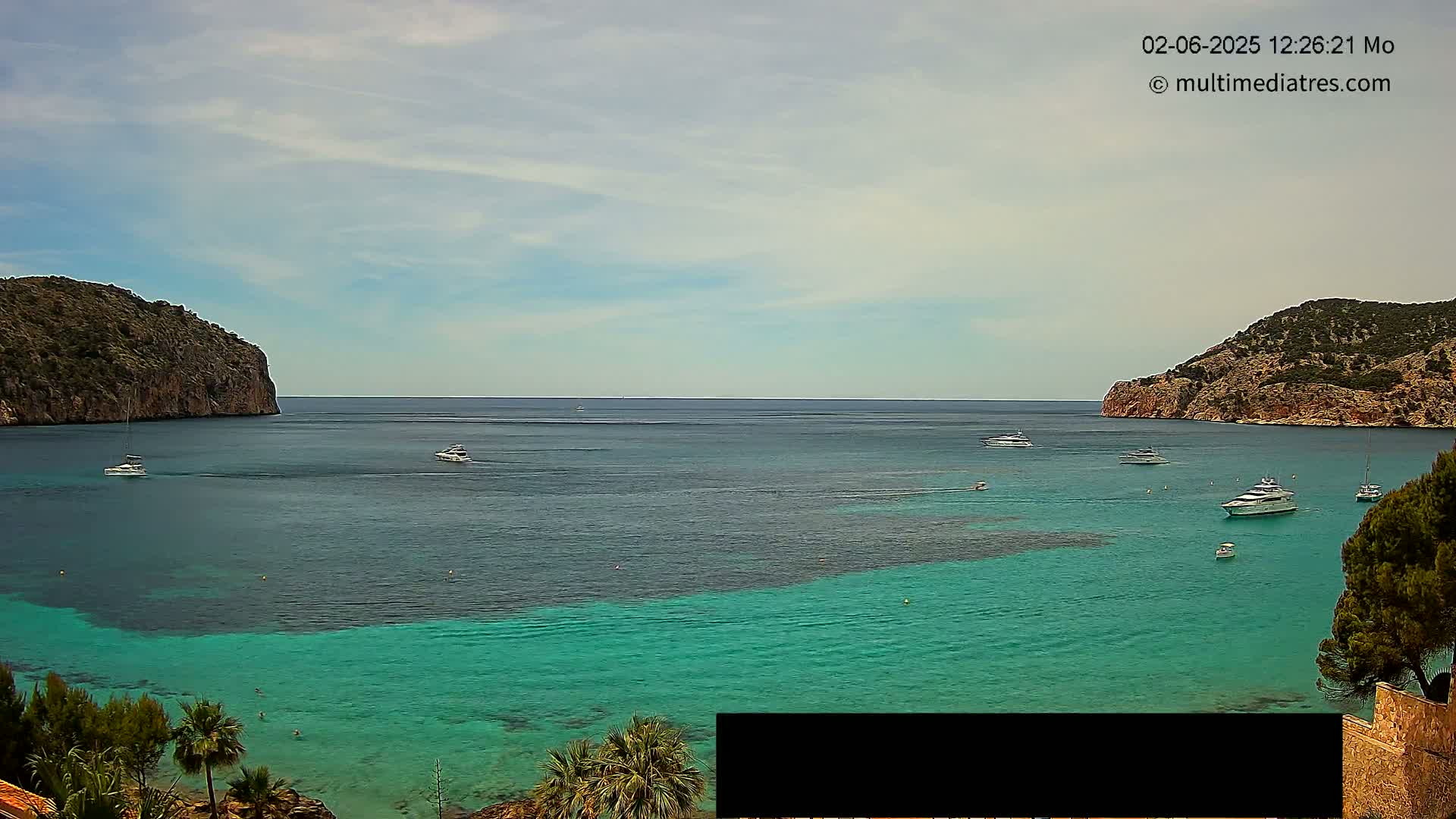 A calm, sunny day reveals a picturesque bay with turquoise water, several boats, and rocky hills on either side.