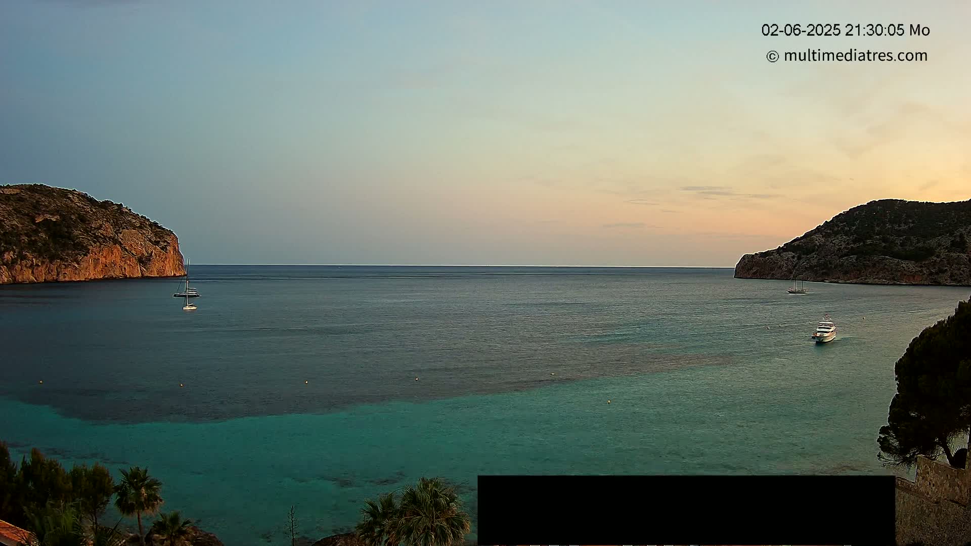 A tranquil, calm ocean cove at sunset is nestled between two rocky hills, with a few boats anchored in the water.