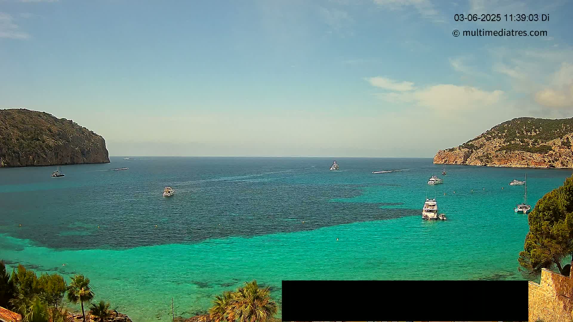 A tranquil cove with clear turquoise water is surrounded by rocky hills under a mostly sunny sky, with several boats scattered across the bay.