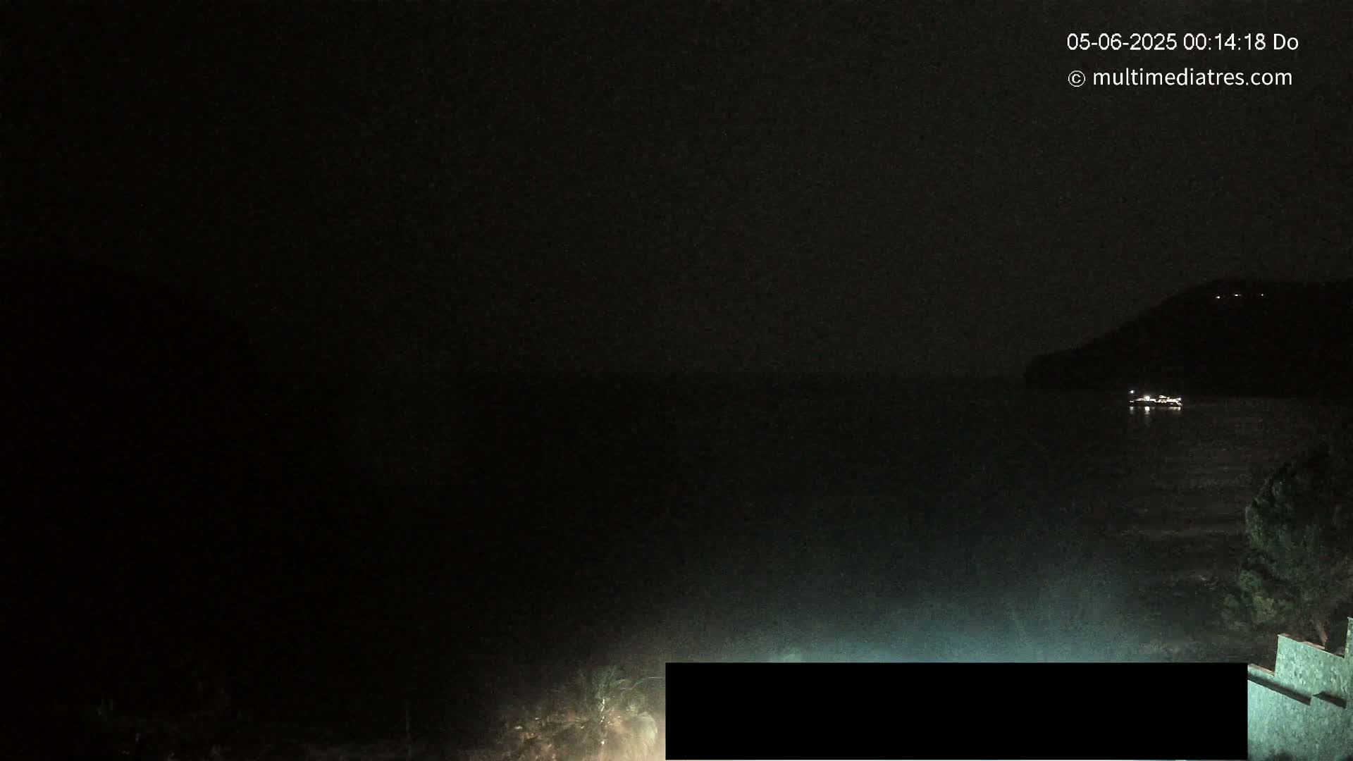 A dark nighttime coastal scene shows a body of water with a distant hill and a few lights visible, under a clear, dark sky.