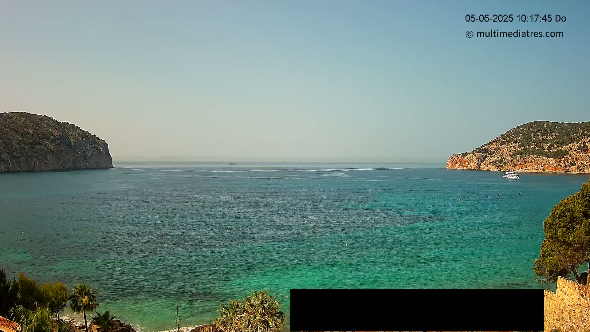 A calm, clear day shows a tranquil bay of teal water, nestled between two rocky hillsides, with a boat visible in the distance.
