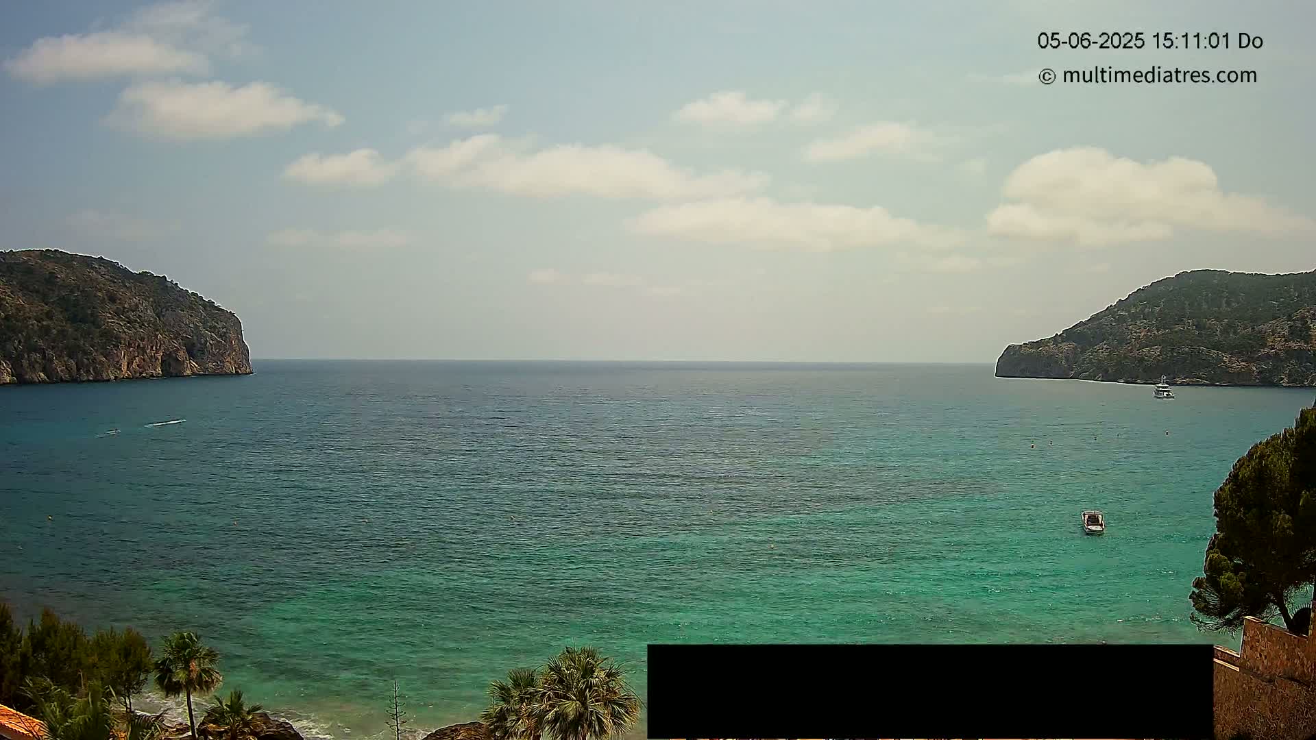 A tranquil bay with clear, teal water is nestled between two rocky hills under a partly cloudy sky.