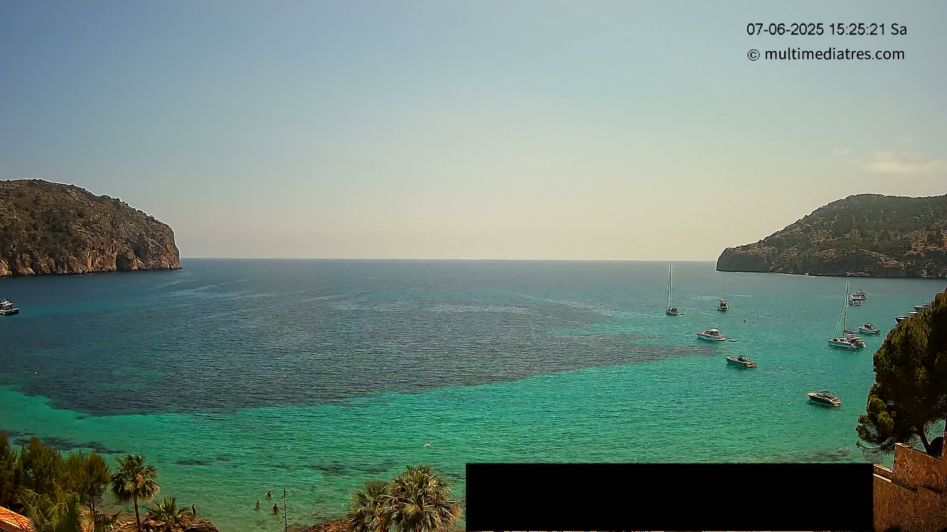 A calm, sunny day reveals a bay of clear, teal water surrounded by rocky hills, dotted with several sailboats and motorboats.