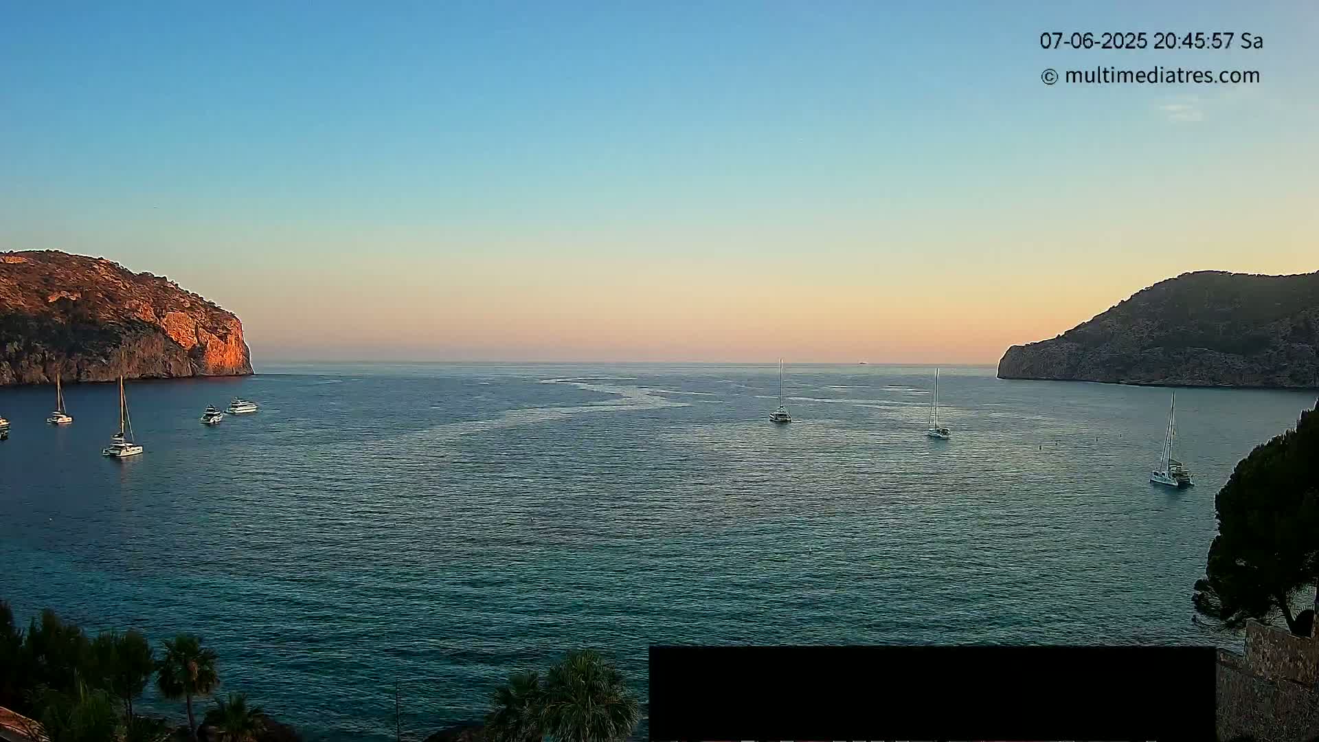 Several boats are anchored in a calm bay between two rocky hillsides at sunset.