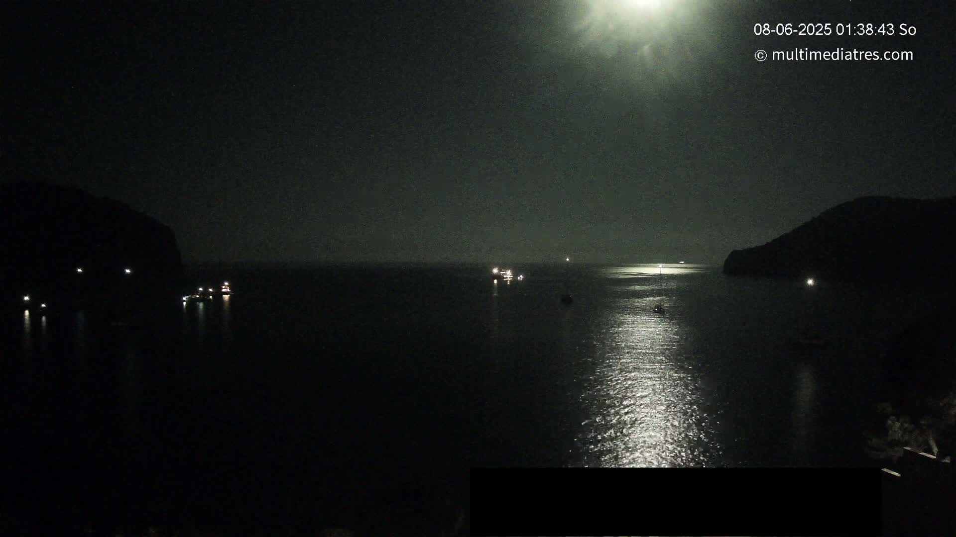 A nighttime view of a calm body of water with several small boats and a bright light source in the sky reflecting on the water's surface, with dark landmasses on either side.