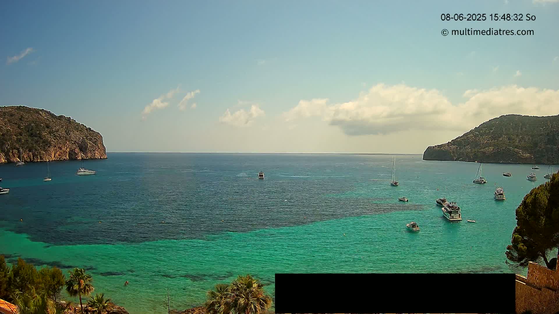 A partly cloudy day shows a calm bay of teal and deep blue water with numerous boats and yachts anchored near rocky cliffs.