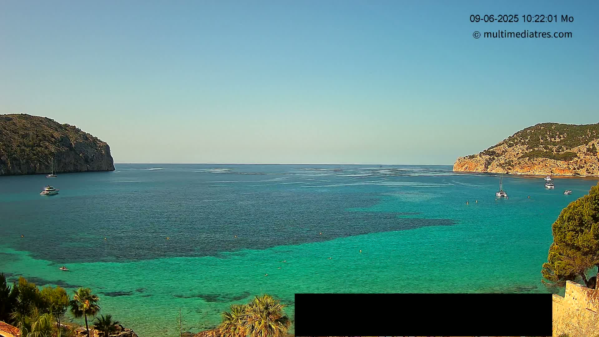 A clear, sunny day reveals a calm bay with teal water, several boats, and rocky hillsides on either side.