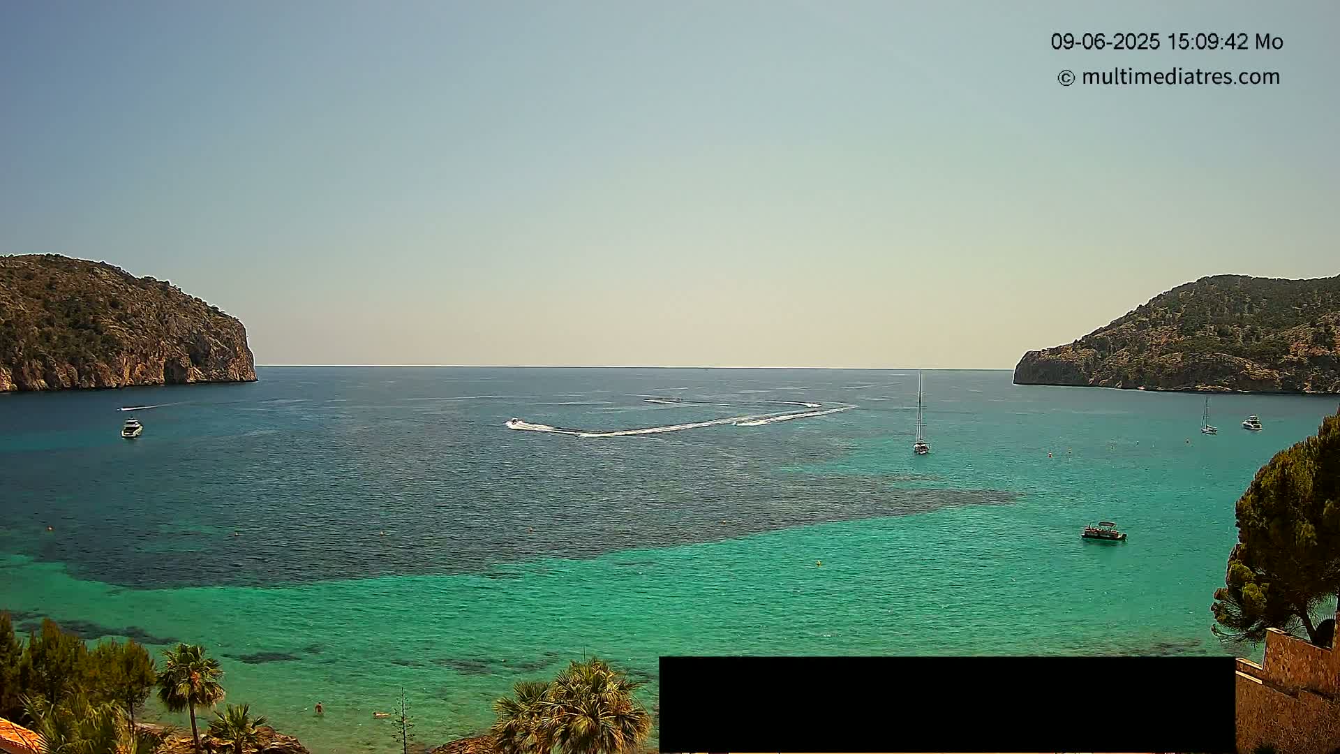 A calm, sunny day reveals a clear turquoise bay surrounded by rocky hills, with several boats and a jet ski trail visible on the water.