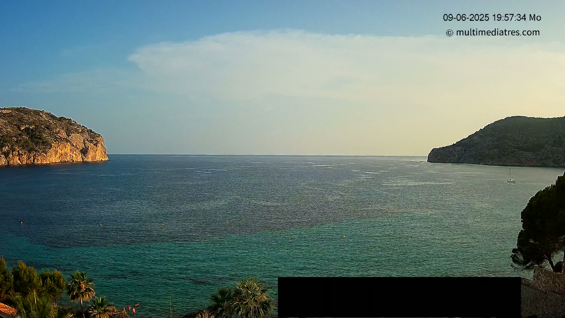 A calm, clear day shows a picturesque coastal scene with two rocky hills flanking a bay of teal and dark-blue water.