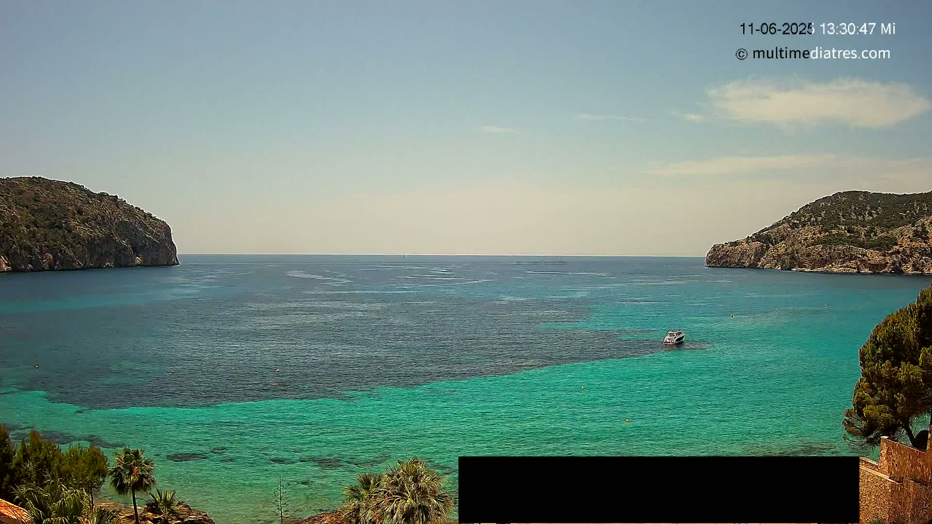 A boat is visible on a calm, bright day in a bay between two rocky hills with clear, teal water.