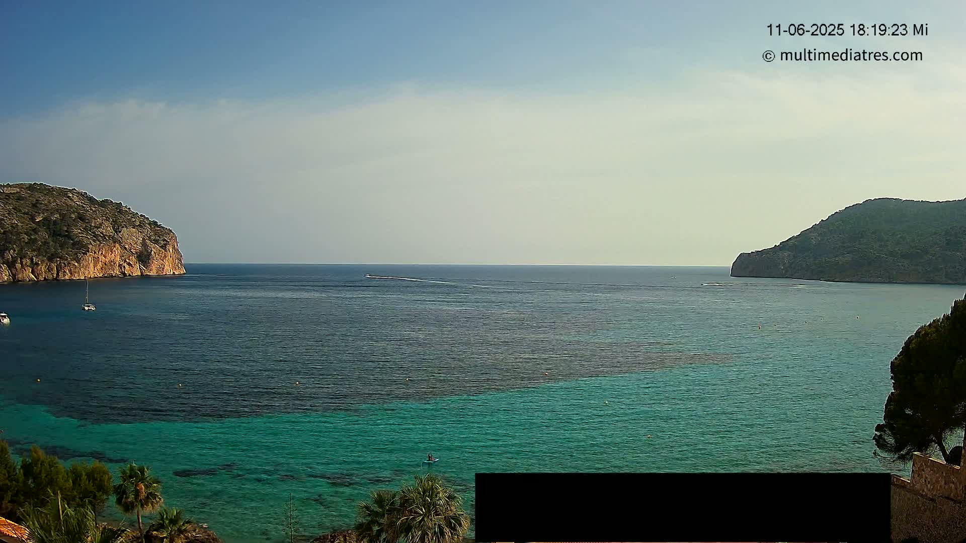 A calm, clear day reveals a picturesque coastal scene with teal-colored water nestled between two rocky hills, a few boats scattered across the bay.