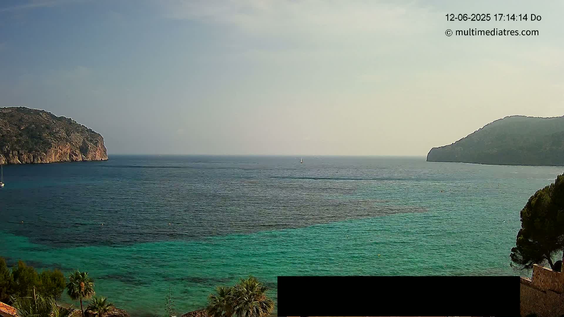 A calm, hazy day shows a tranquil bay of teal and dark blue water, enclosed by two rocky hills, with a lone sailboat in the distance.