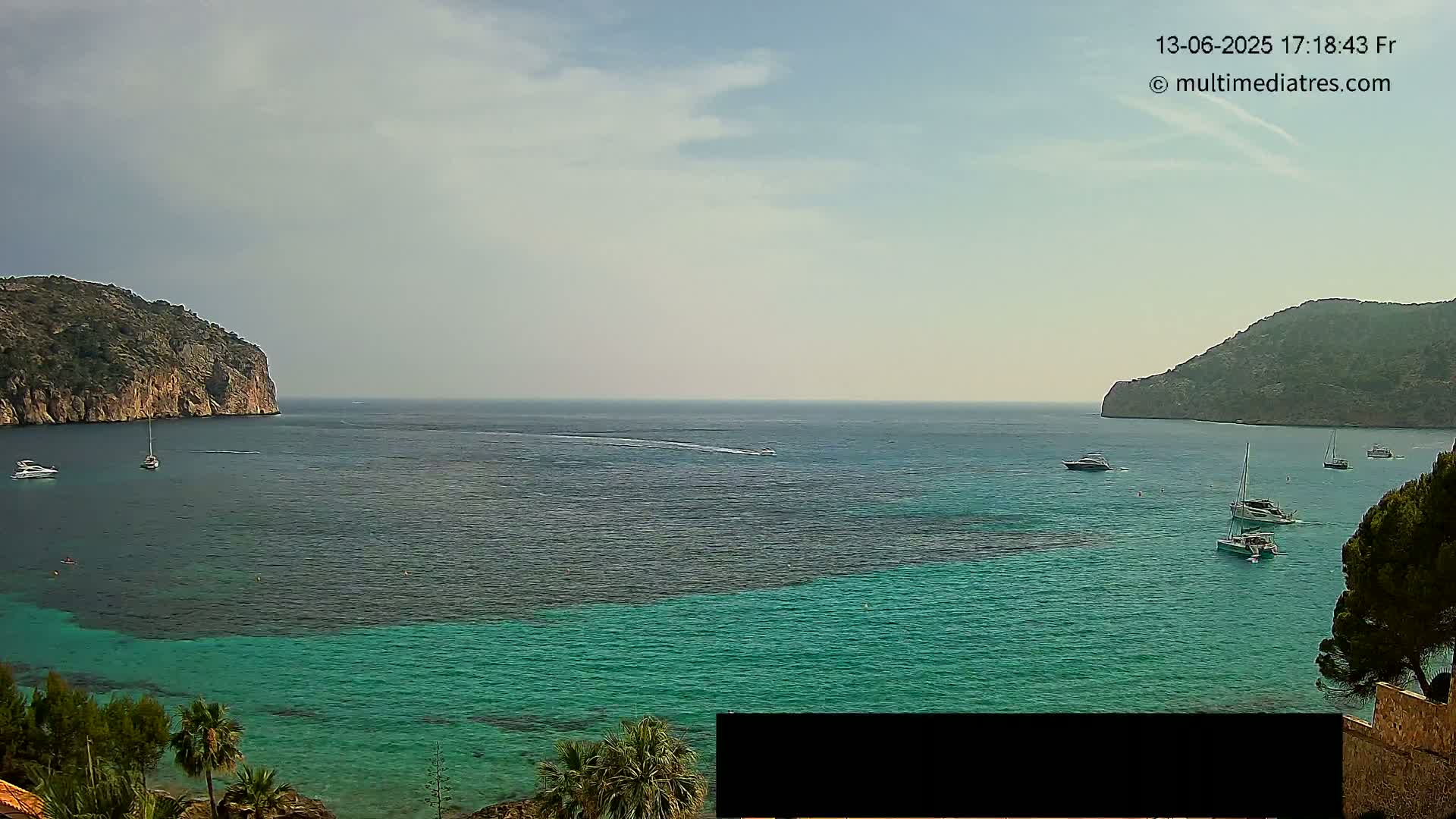 A calm, partly cloudy day reveals a picturesque bay with teal water, several boats, and rocky hillsides on either side.