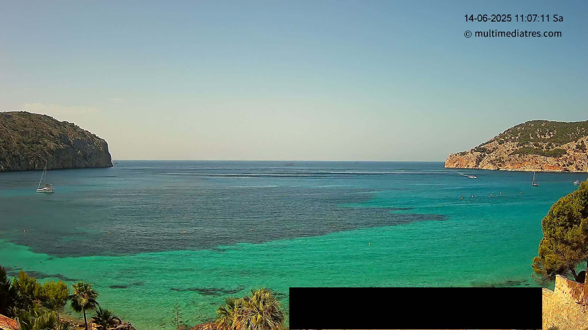 A tranquil bay with clear, turquoise water is nestled between two rocky hills under a clear, sunny sky, with several sailboats visible.