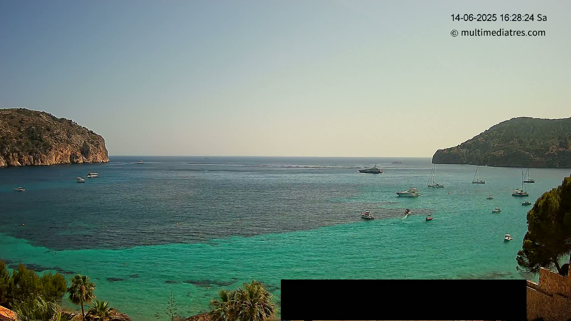A clear, sunny day reveals a tranquil bay with numerous boats anchored in vibrant turquoise water, surrounded by rocky hillsides.