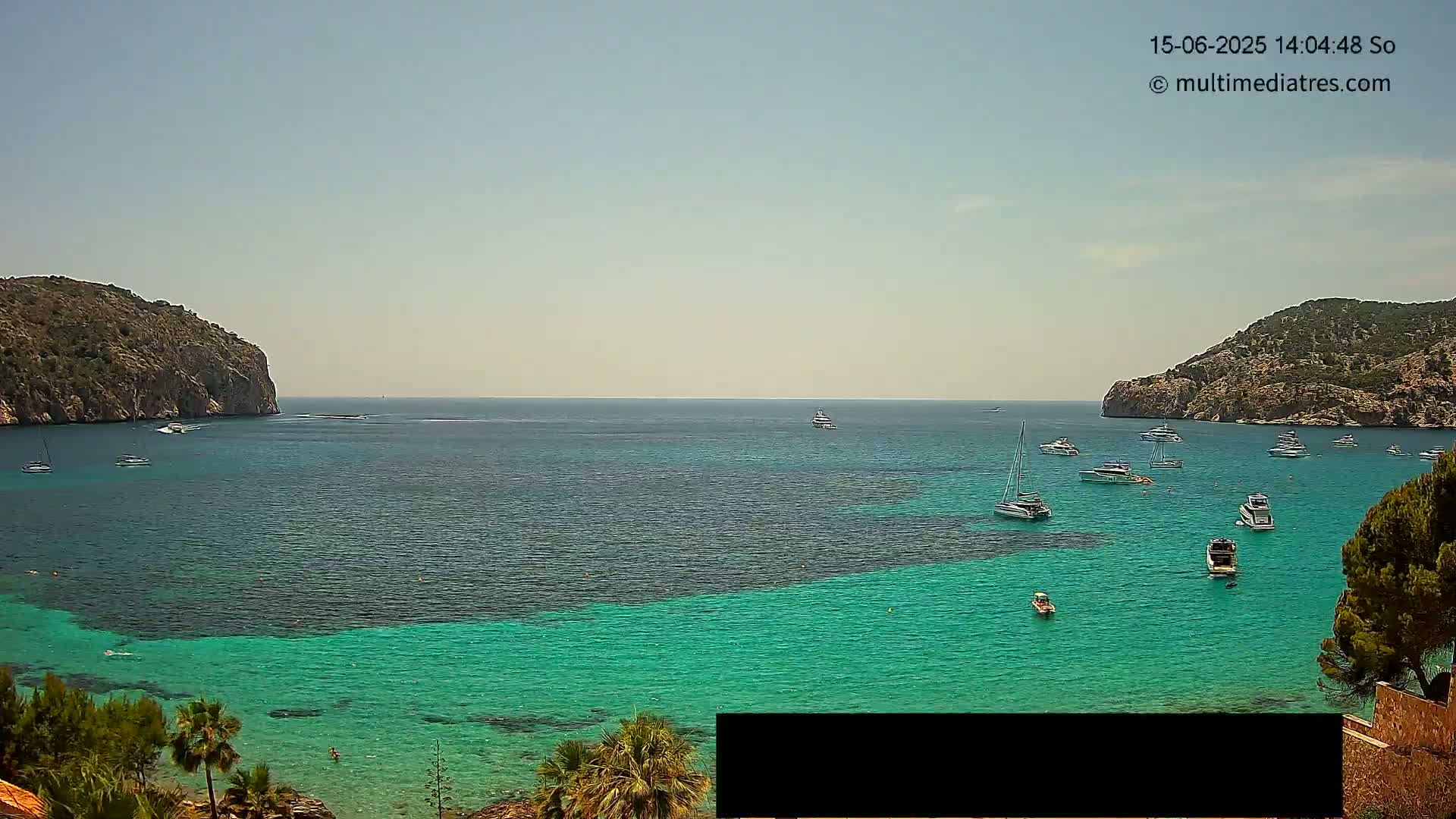 A calm, sunny day reveals a picturesque bay with turquoise water, numerous boats, and two rocky hillsides on either side.