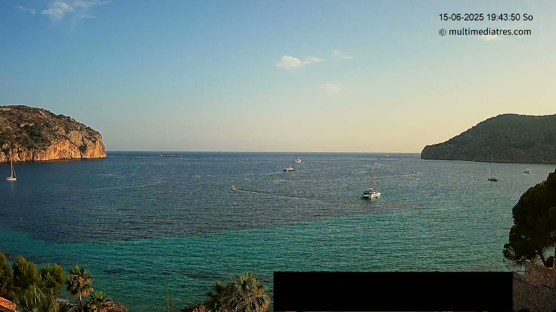 Under a clear, mostly sunny sky, a calm bay is dotted with several boats near two rocky hillsides.