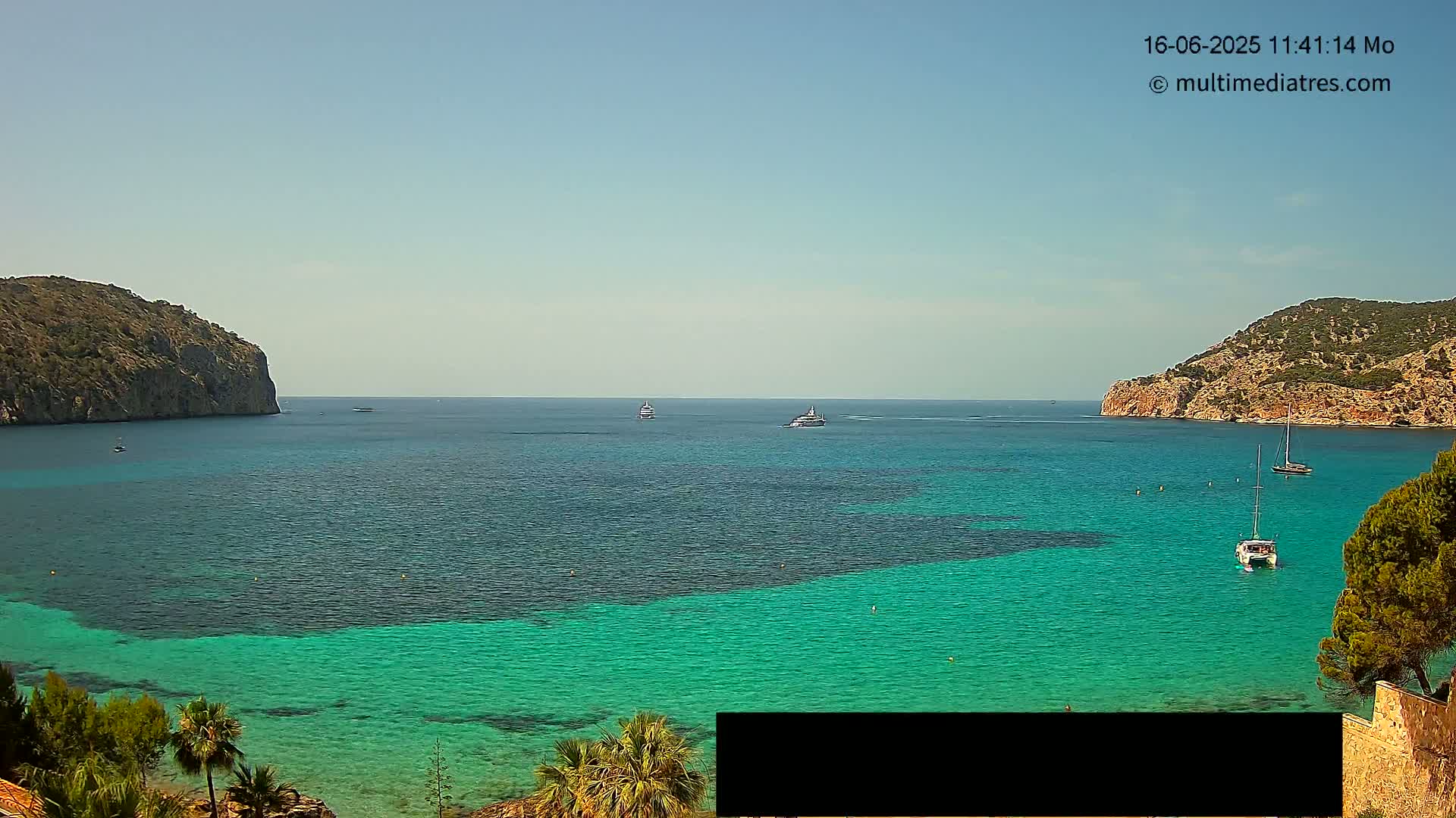 A clear, sunny day reveals a calm, turquoise bay nestled between two rocky hills, with several boats scattered across the water.
