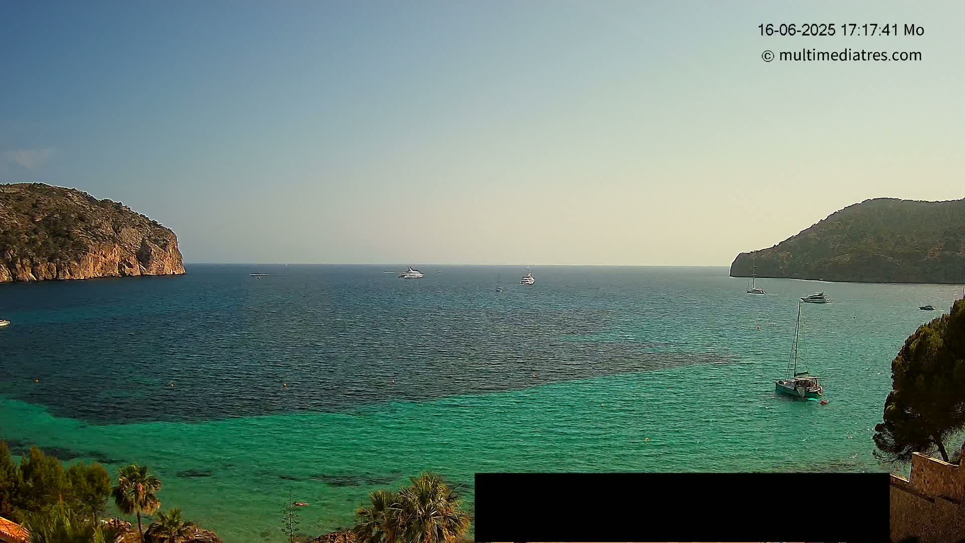A tranquil bay with clear turquoise water is nestled between two rocky hills under a bright, sunny sky, with several boats scattered across the water.