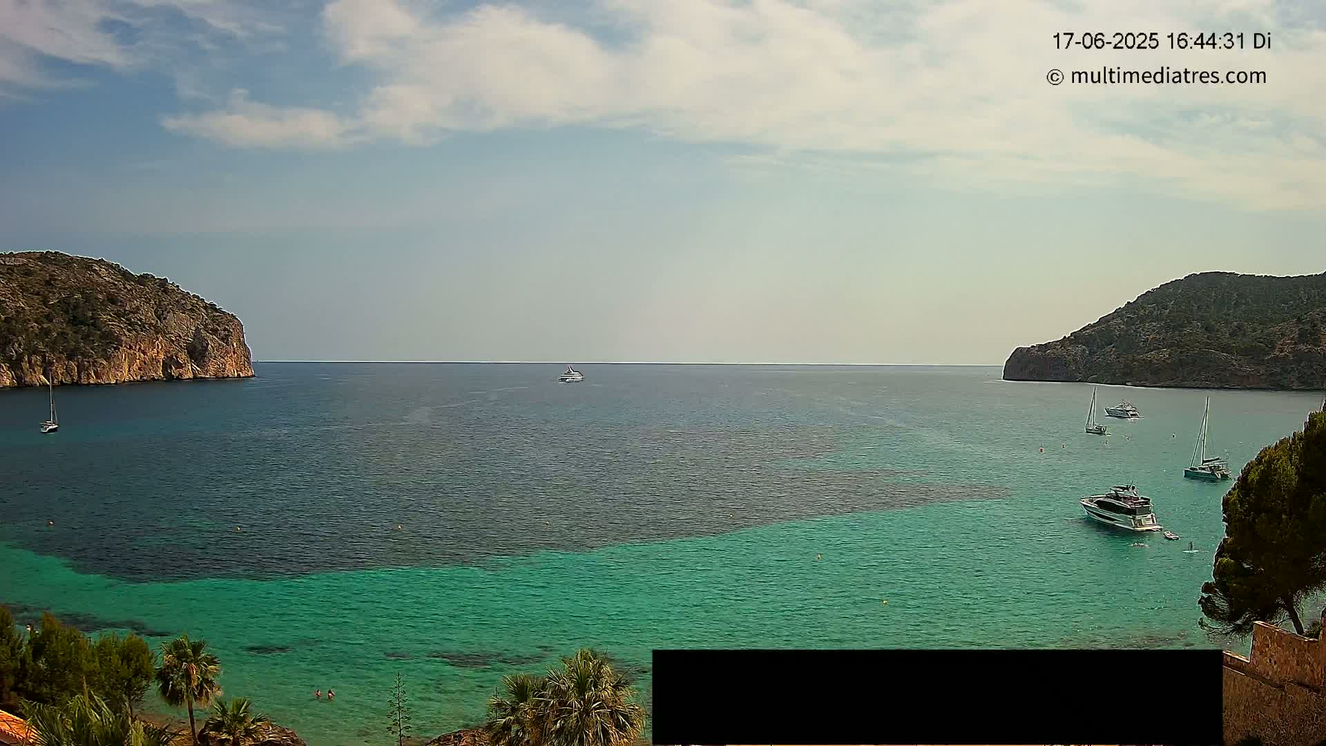 A partly cloudy day shows a calm bay of teal and dark-blue water between two rocky hills, with several boats and yachts scattered across the water.