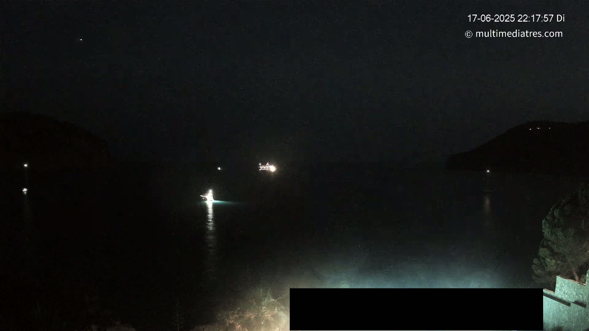 At night, several boats with lights are visible on a dark body of water between two darkened landmasses.