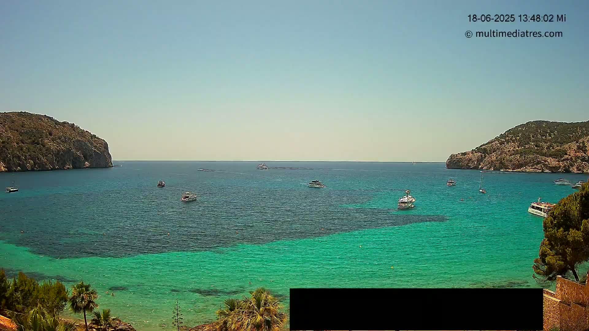 A calm, sunny day shows a bay of bright turquoise water with several boats, surrounded by rocky hills and vegetation.