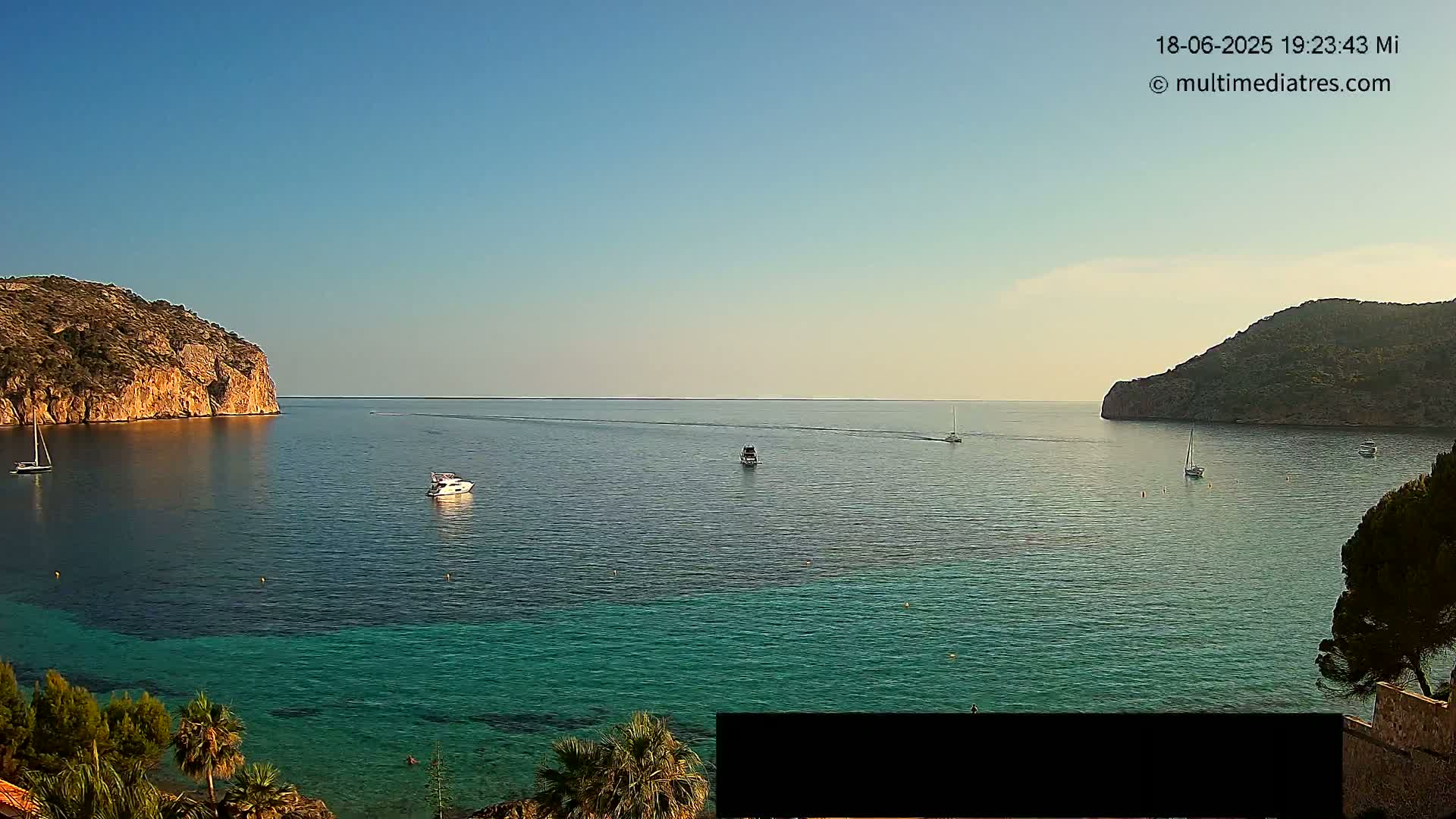 A calm, clear day reveals a tranquil bay with several boats, surrounded by rocky hillsides and lush greenery.