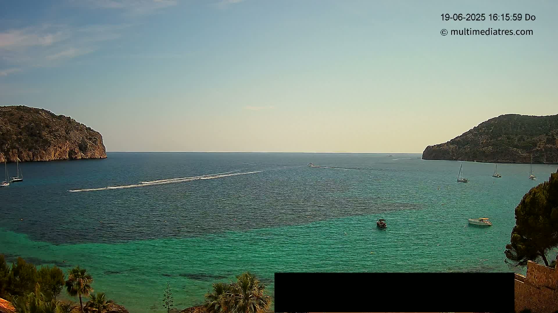 A tranquil bay with clear turquoise water is surrounded by rocky hills under a mostly clear sky, with several boats scattered across the water.