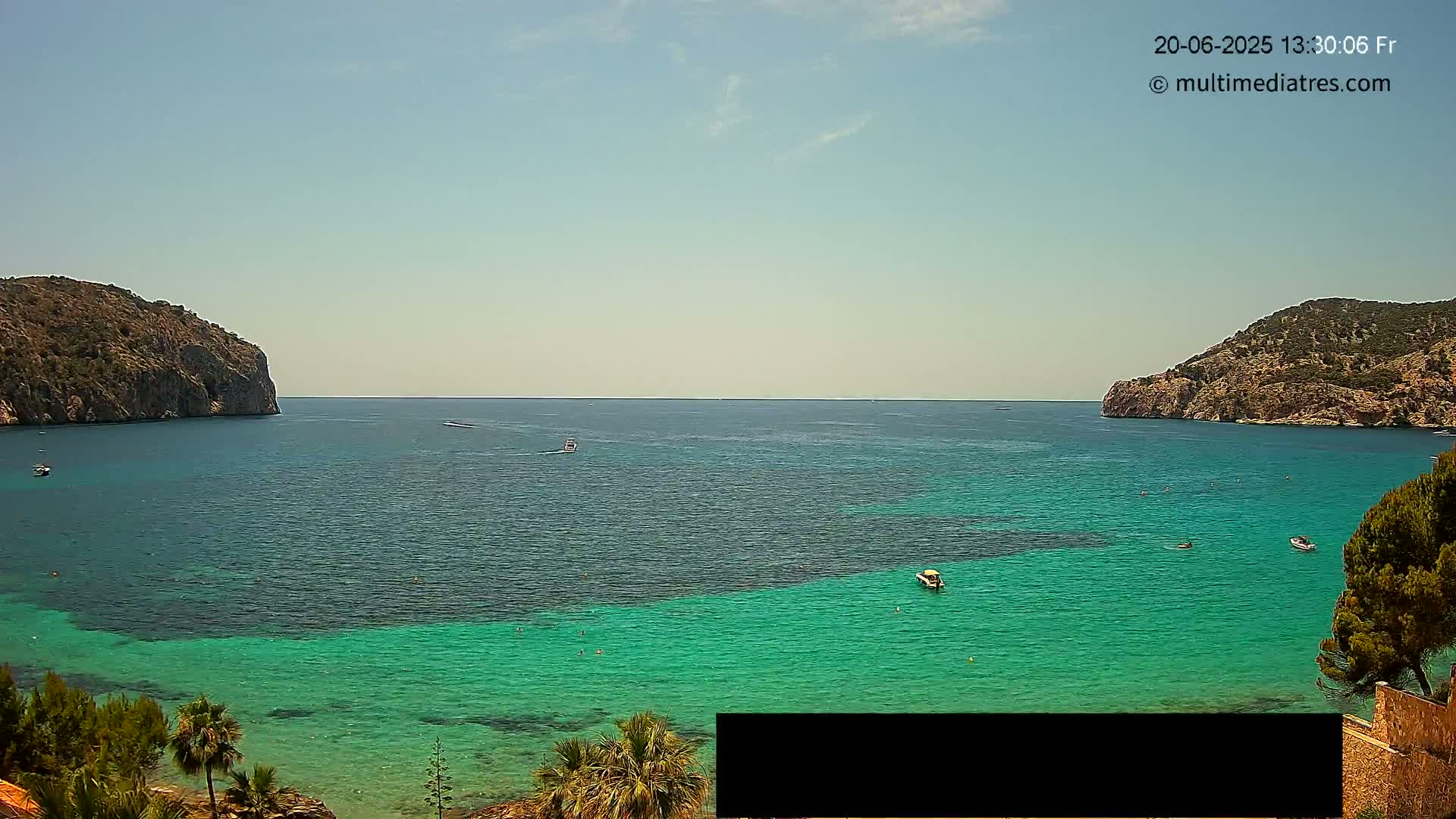 A clear, sunny day shows a calm, teal-colored bay nestled between two rocky hillsides, with several small boats scattered across the water.