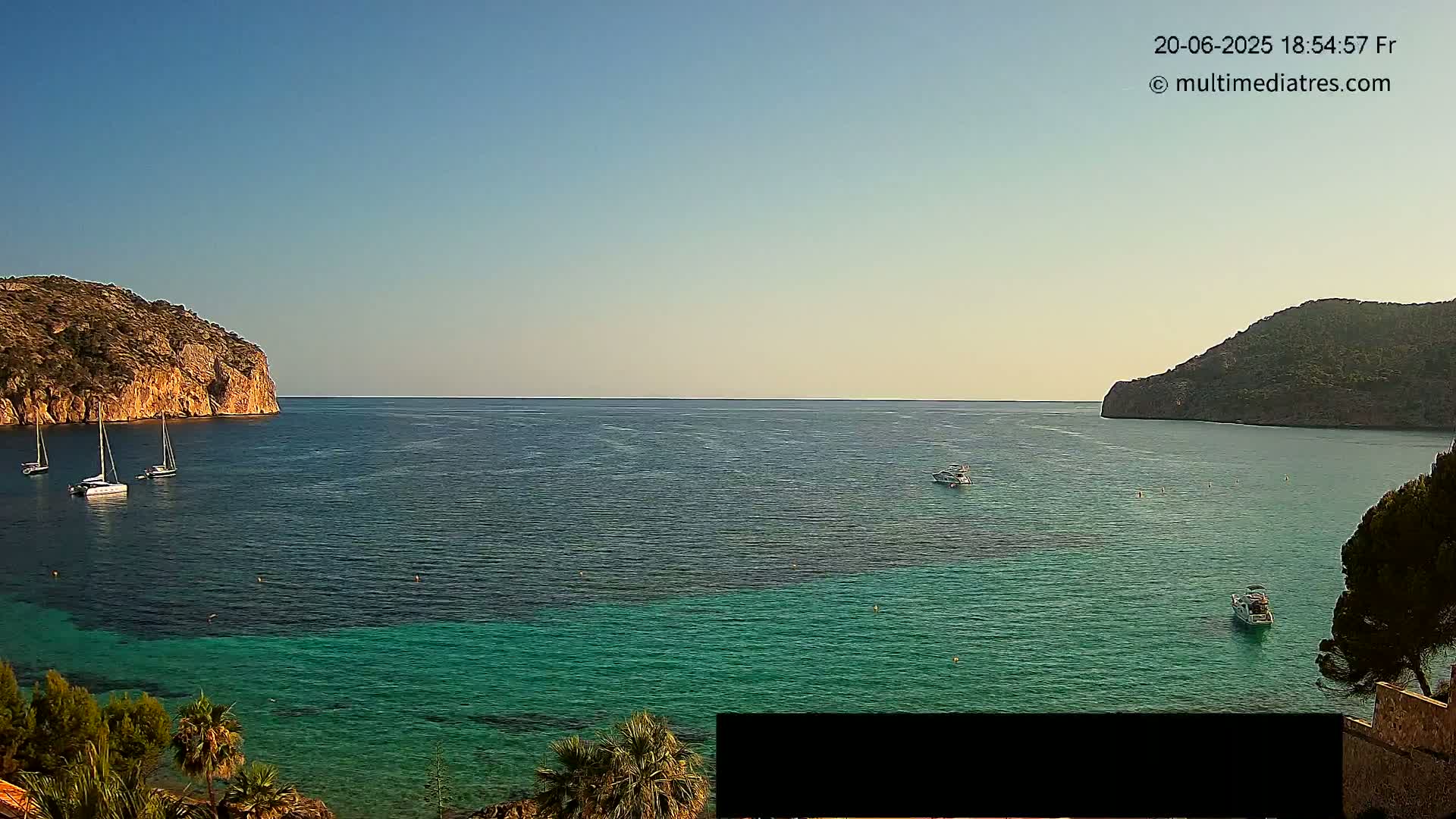 A tranquil bay with clear, teal water is surrounded by rocky hills under a clear, bright sky; several sailboats and small motorboats are visible on the water.