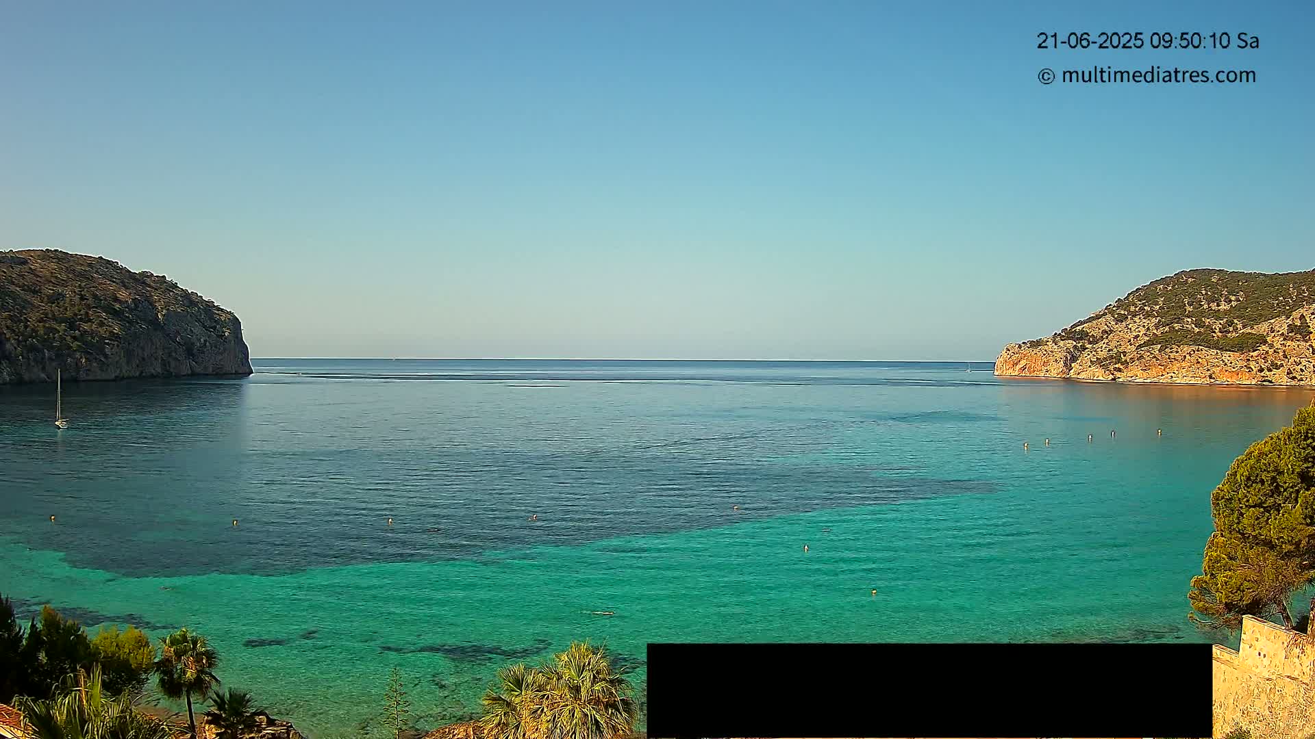 A clear, sunny day reveals a calm bay of teal water nestled between two rocky hillsides, with a small sailboat visible in the distance.