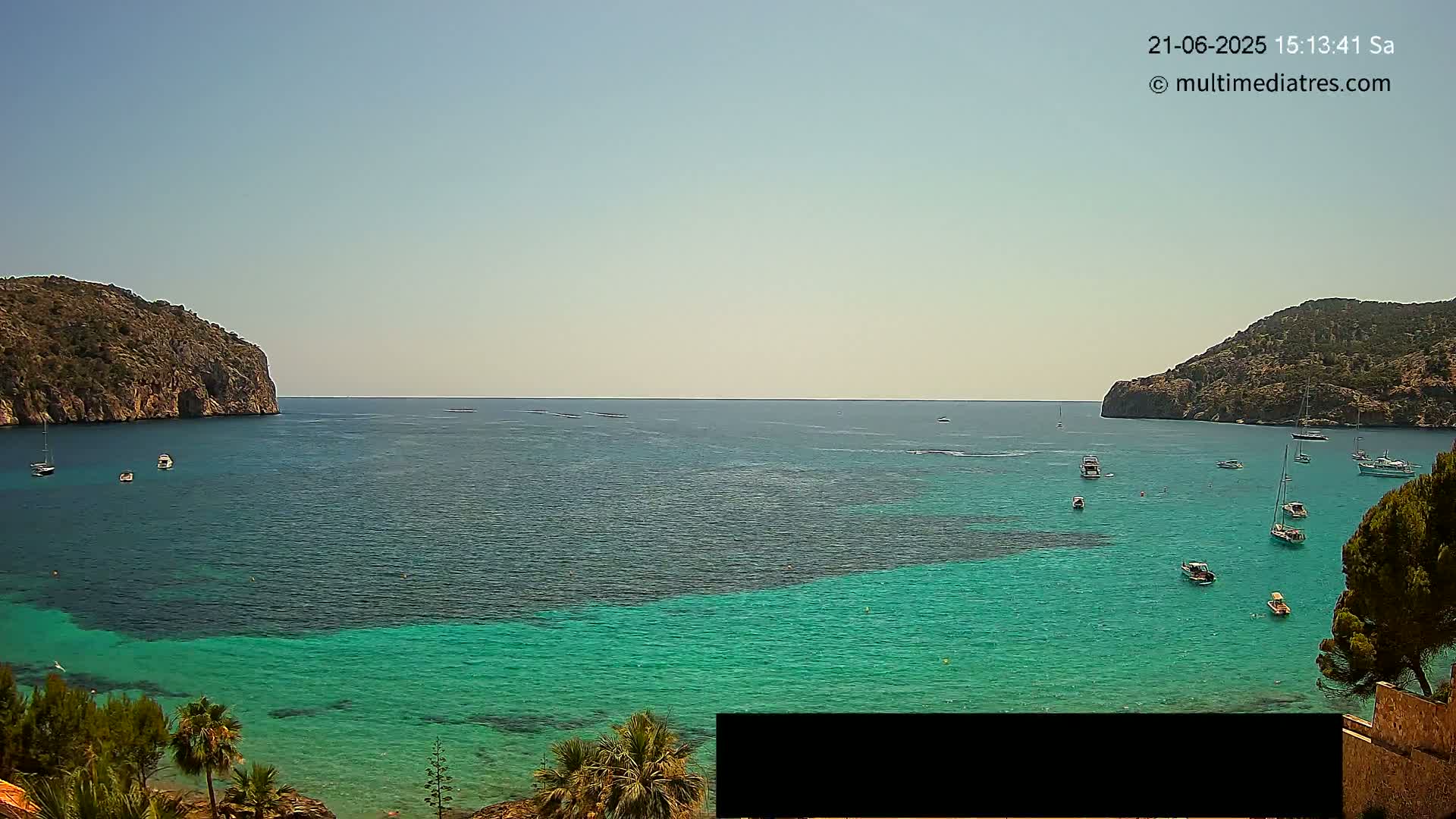 A calm, sunny day reveals a bay of turquoise water, dotted with sailboats, nestled between two rocky hillsides.