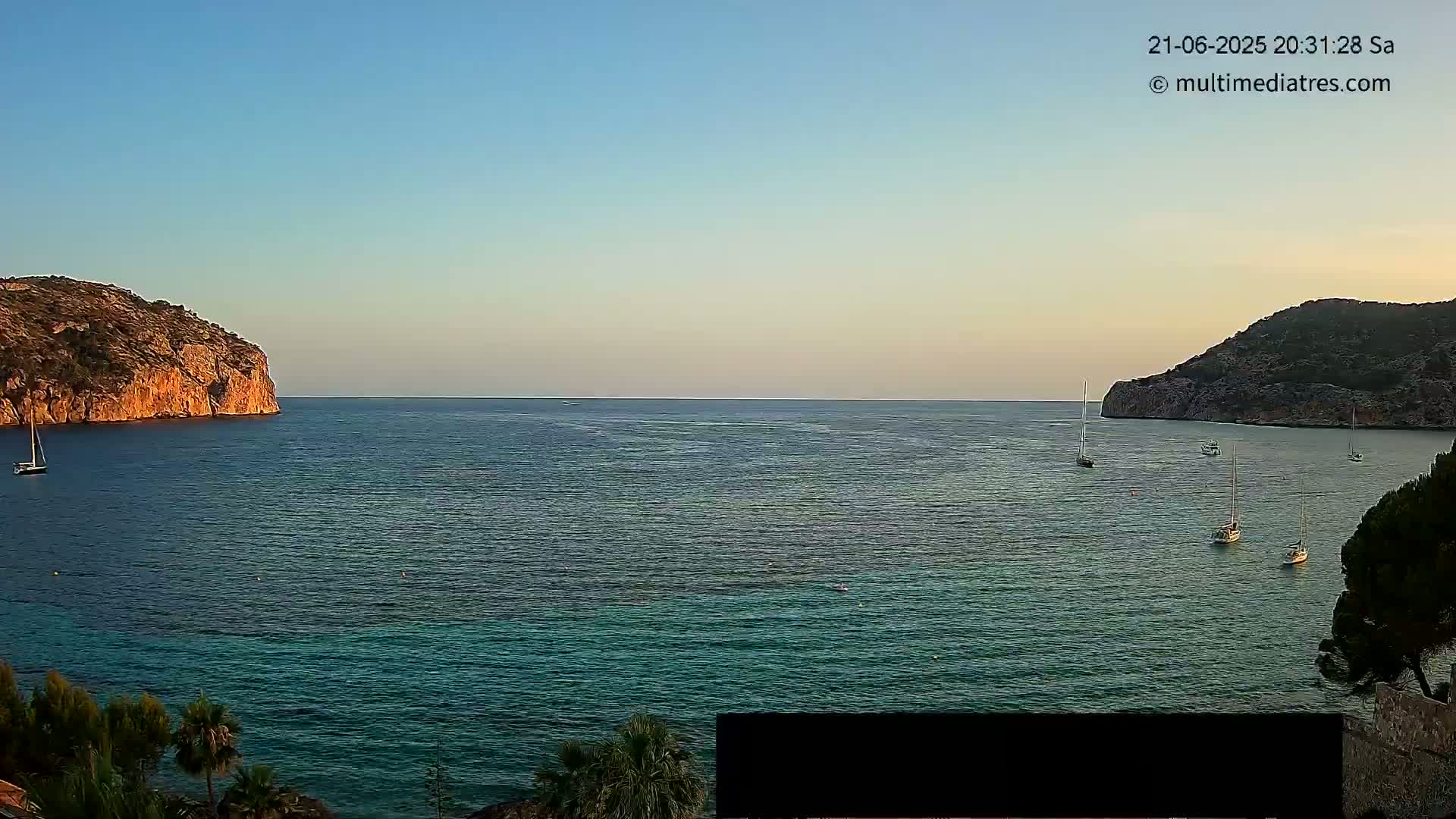 Several sailboats are anchored in a calm, teal-colored bay between two rocky hills under a clear, twilight sky.