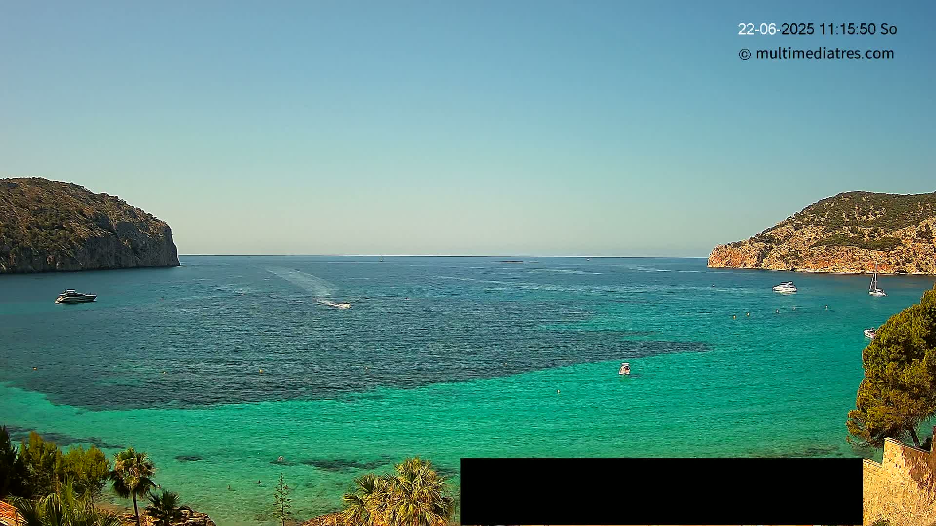 A tranquil cove with clear, turquoise water is nestled between two rocky hills on a sunny day, with several boats scattered across the water.
