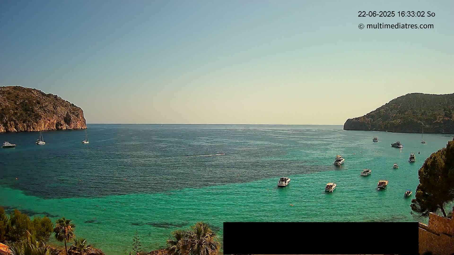 Under a clear blue sky, a calm bay is dotted with numerous boats and yachts, nestled between two rocky cliffs.