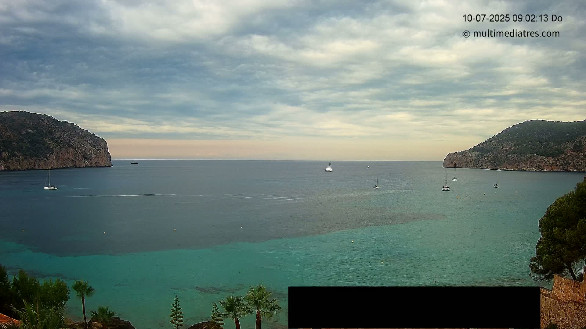 A calm bay with several sailboats, partially obscured by a cloudy sky, is nestled between two rocky hillsides with lush vegetation.